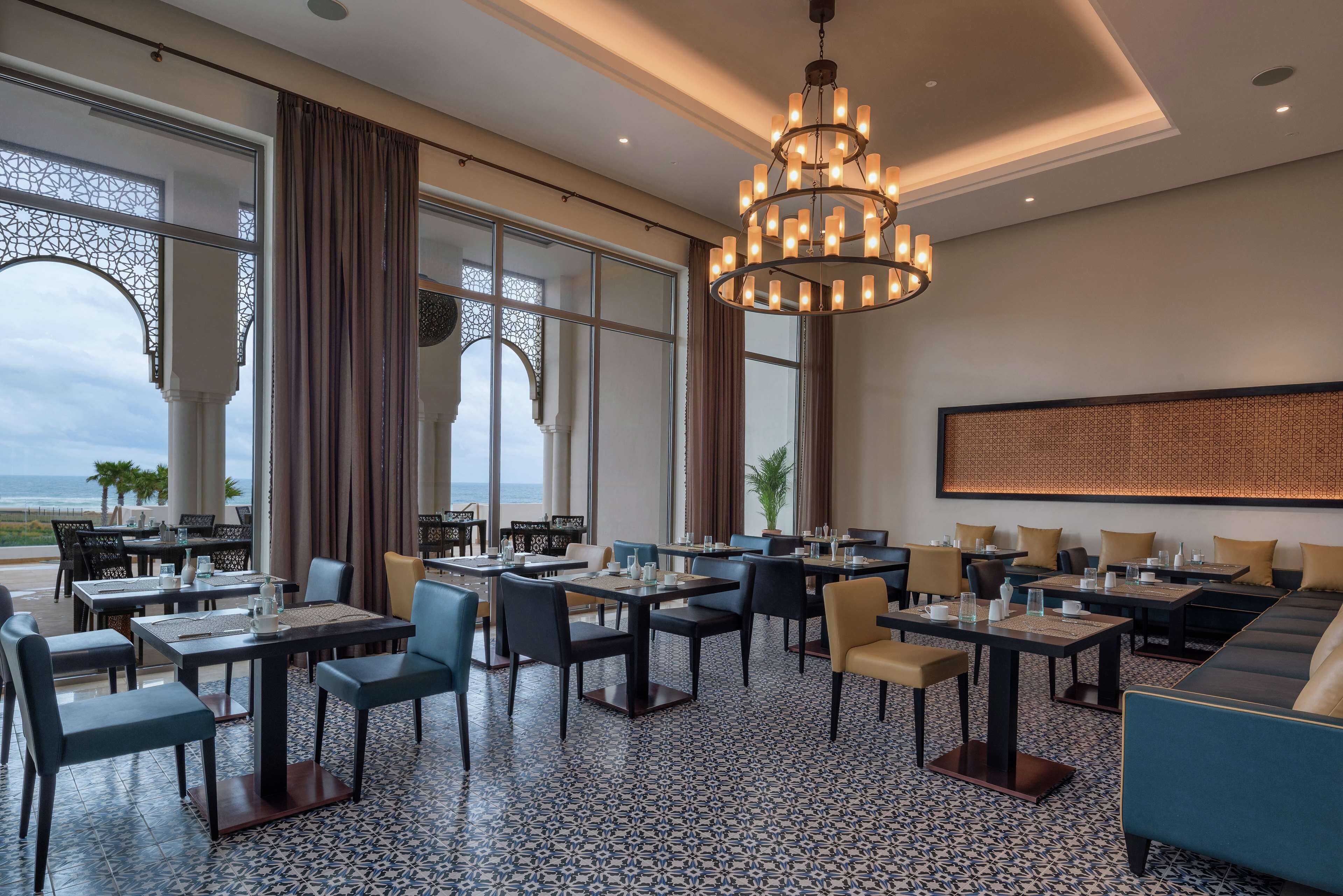 A hotel dining area with tables and chairs, a chandelier, and large windows with curtains.
