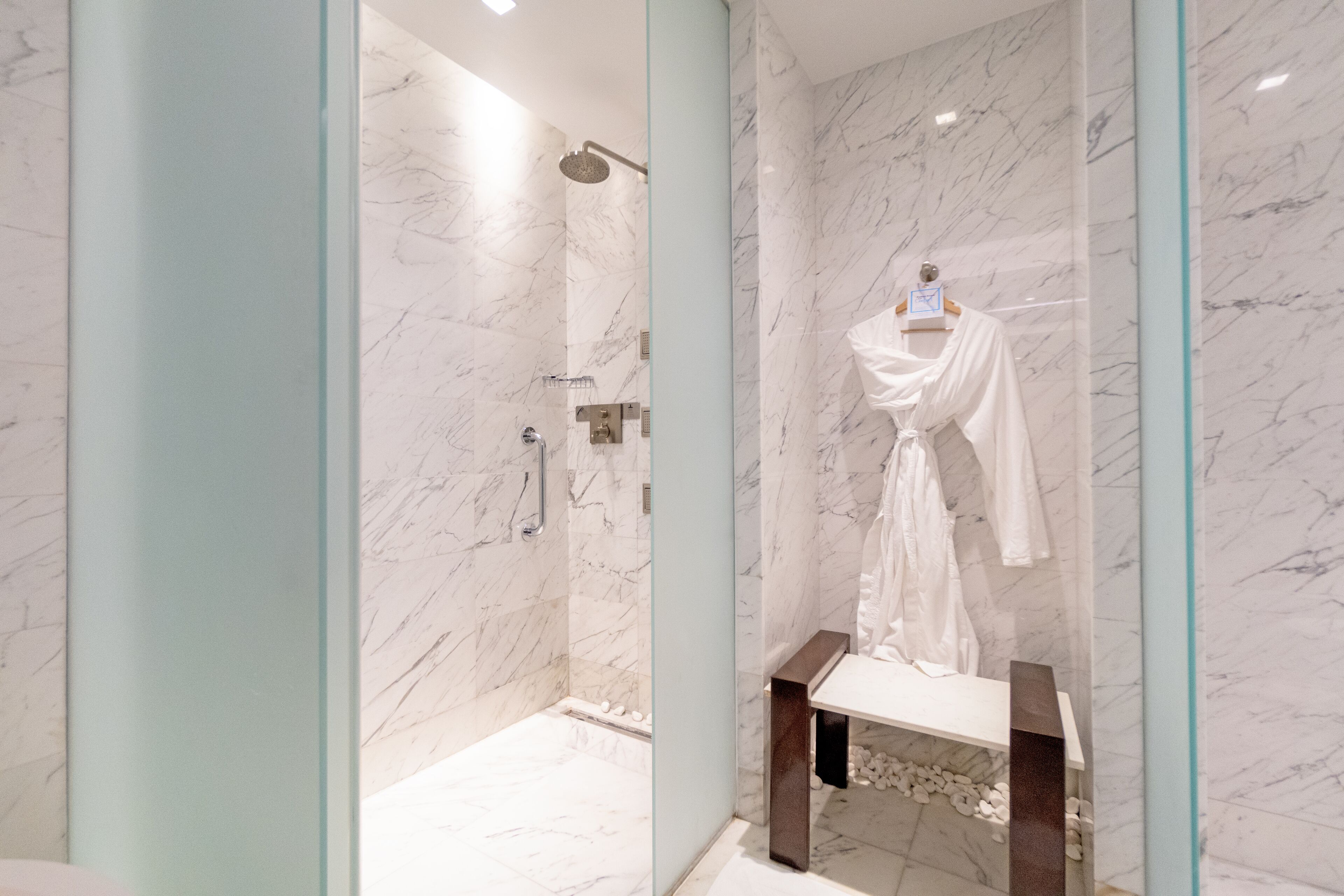 A modern bathroom with a marble shower, a glass-enclosed vanity, and a robe hanging on a rack.