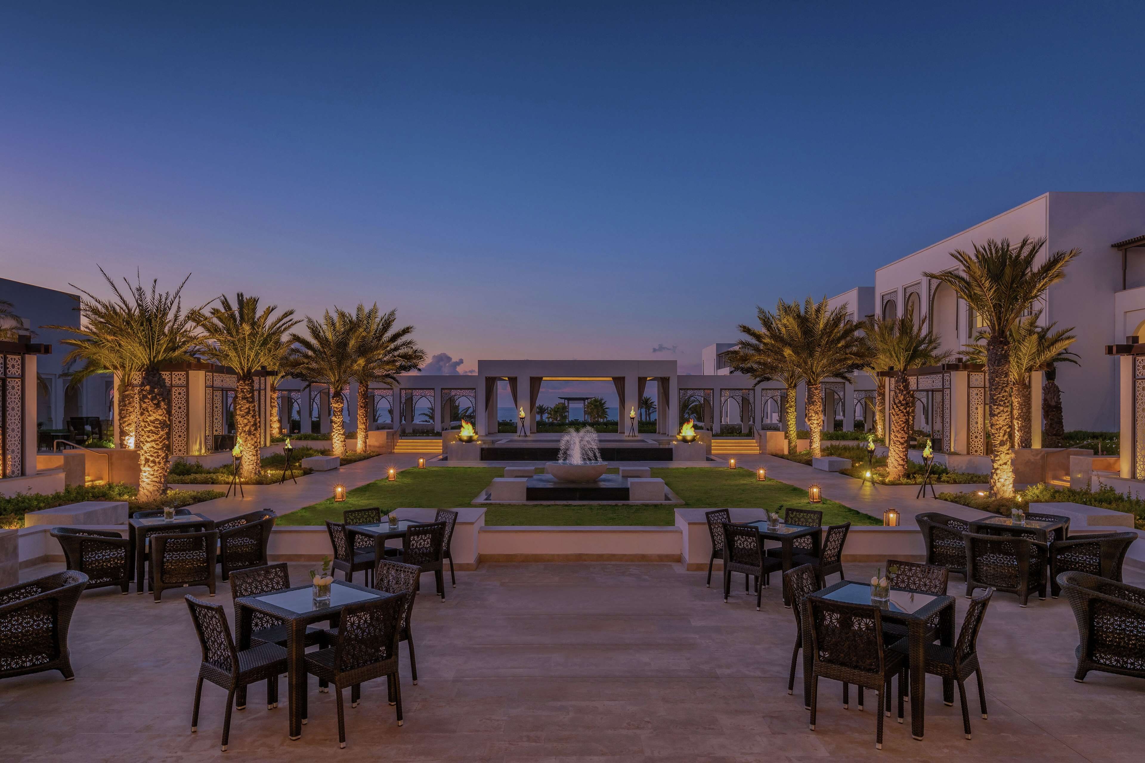 An outdoor dining area with tables and chairs, palm trees, and a water feature.