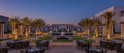 An outdoor dining area with tables and chairs, palm trees, and a water feature.