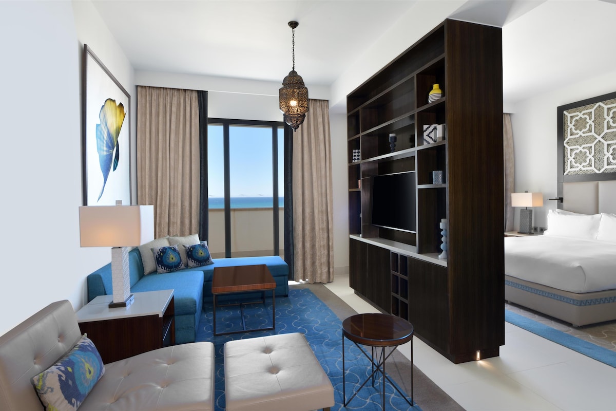 Quarto de hotel moderno com uma cama grande, um sofá, uma mesa de centro, uma TV e uma estante de livros.