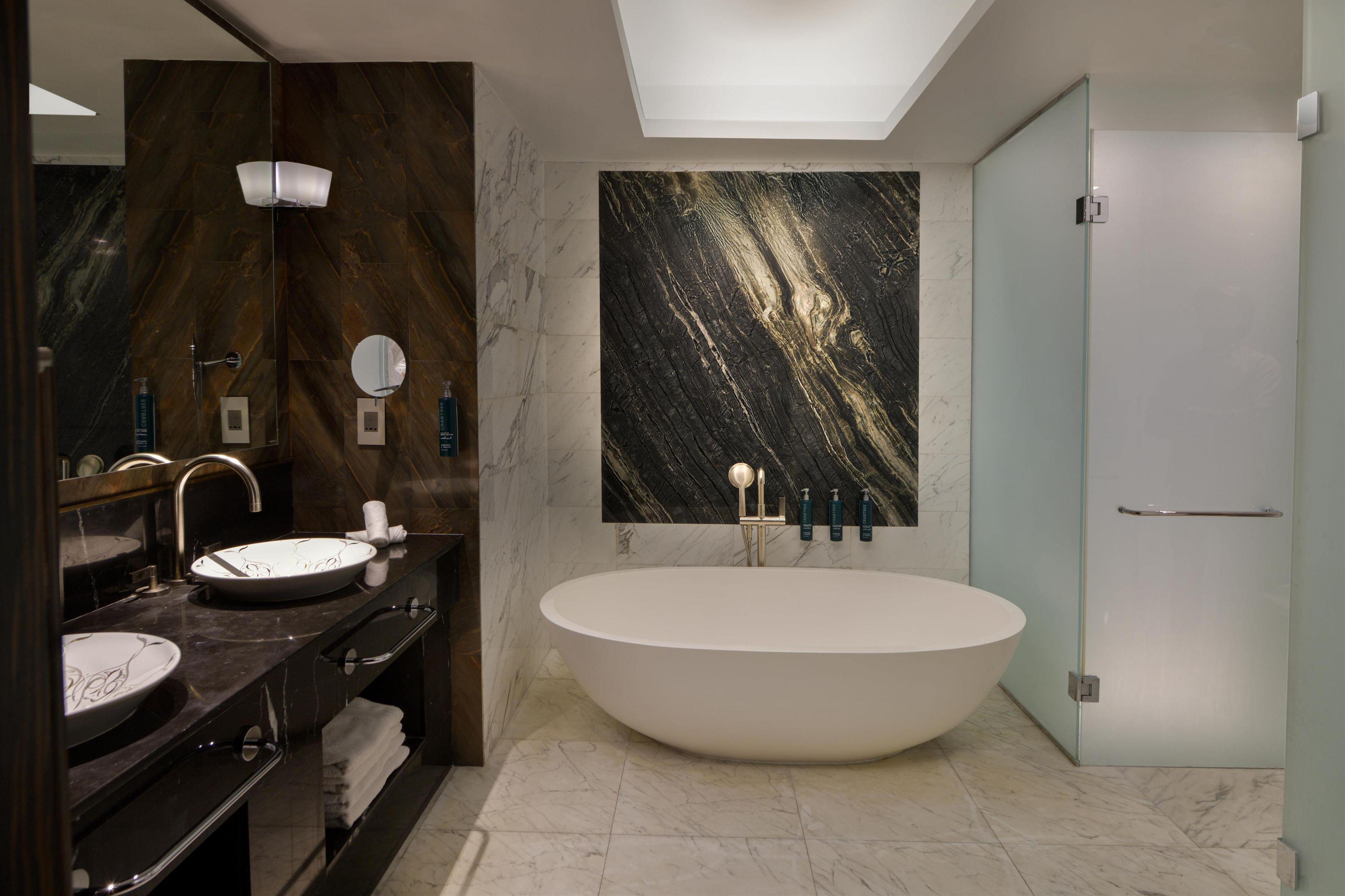 Ein modernes Badezimmer mit freistehender Badewanne, dunklem Holz-Waschtisch und einer Marmor-Spritzschutzwand.