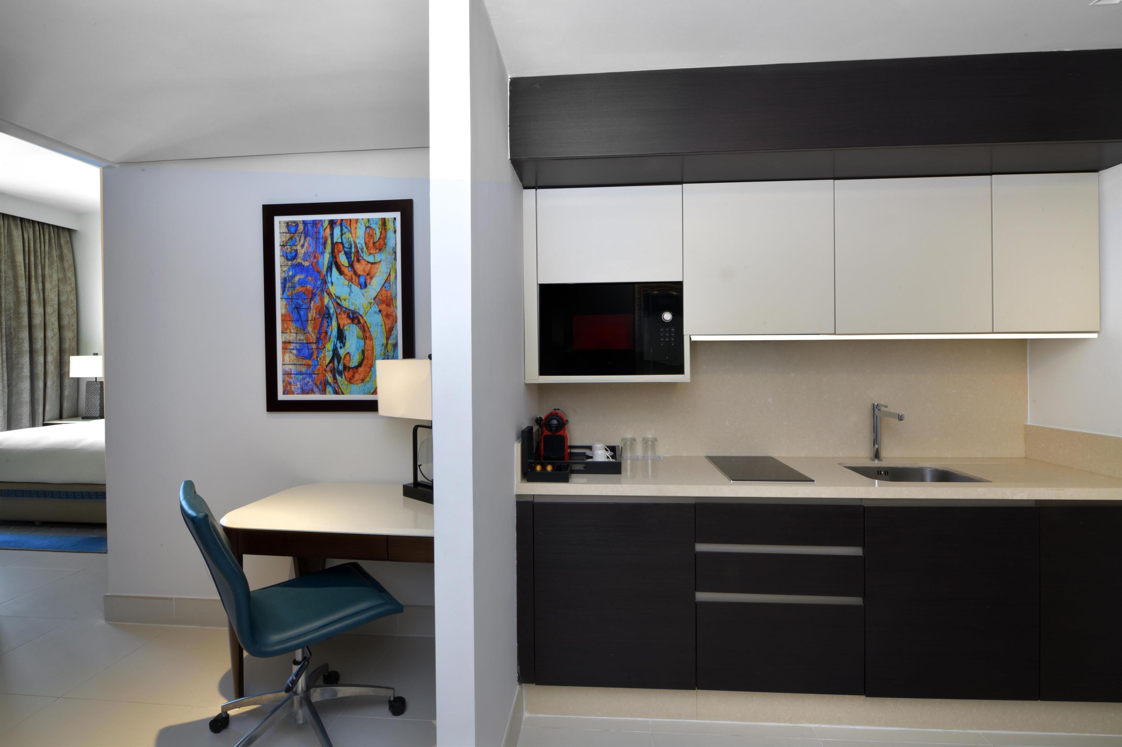 A modern hotel room with a kitchenette, a dining area with a blue chair, a framed abstract painting, and a bed with a grey curtain.