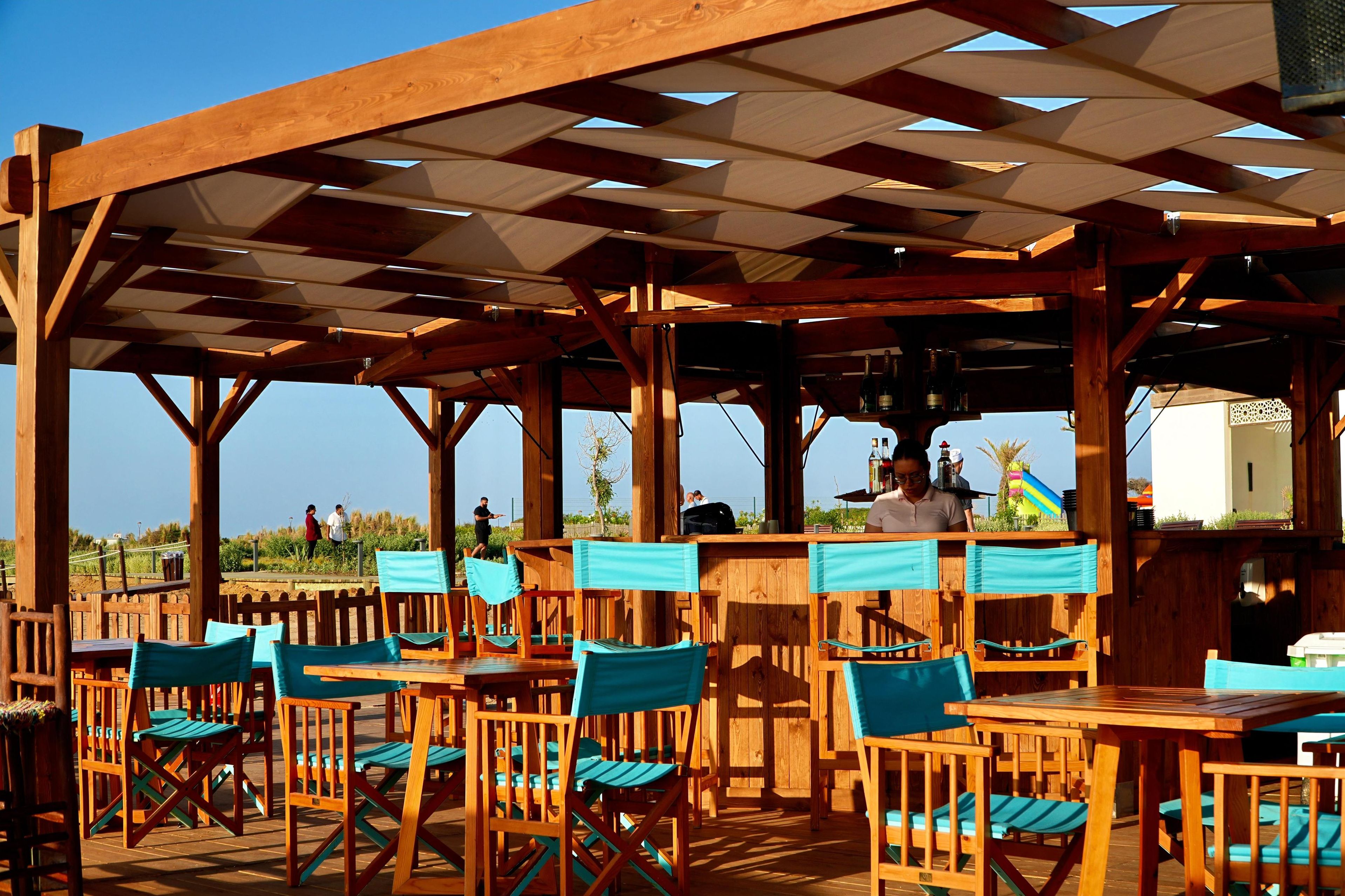 Una terraza de madera al aire libre con mesas y sillas, una zona de bar y vistas a la playa.