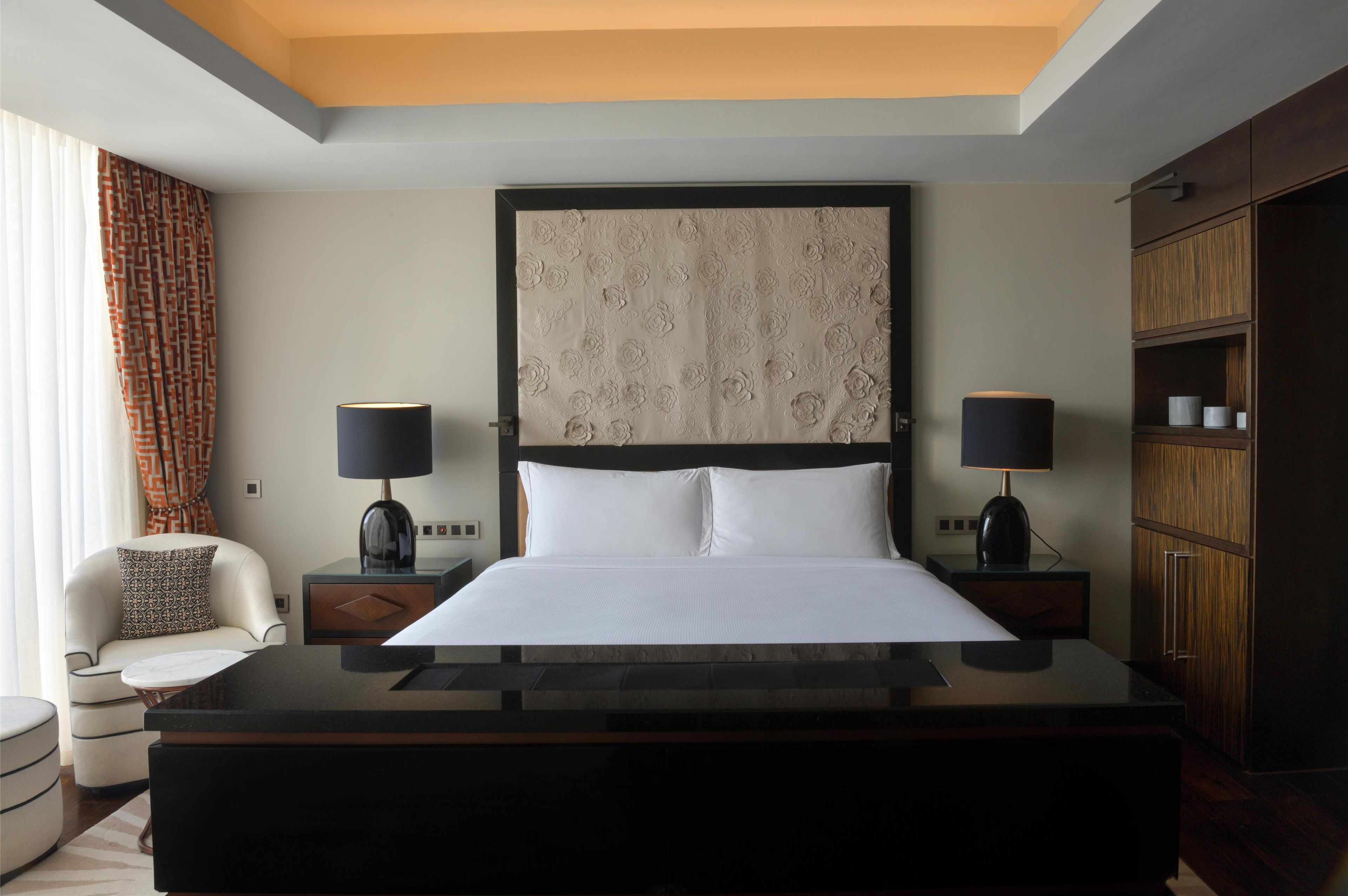 A modern hotel room with a large bed, two bedside tables with lamps, a wooden wardrobe, and a patterned curtain.