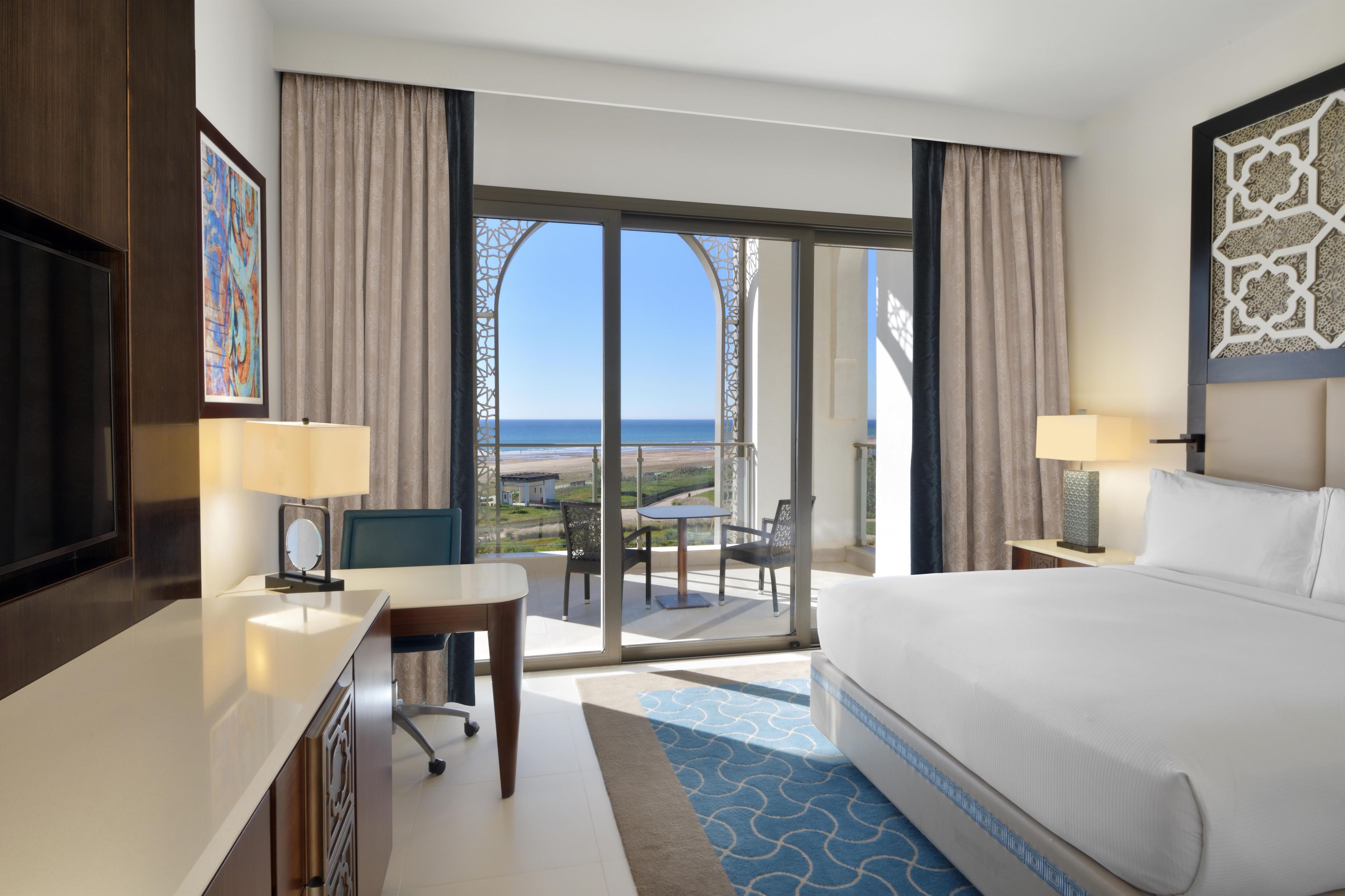 A hotel room with a large bed, a desk, a chair, and a view of the ocean through a glass door.