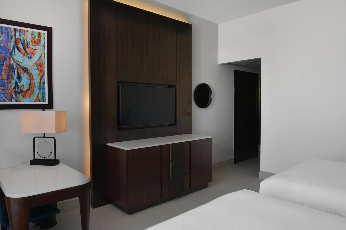 Une chambre d’hôtel moderne avec un téléviseur à écran plat fixé sur un panneau en bois foncé, une table de chevet avec une lampe et un lit blanc.