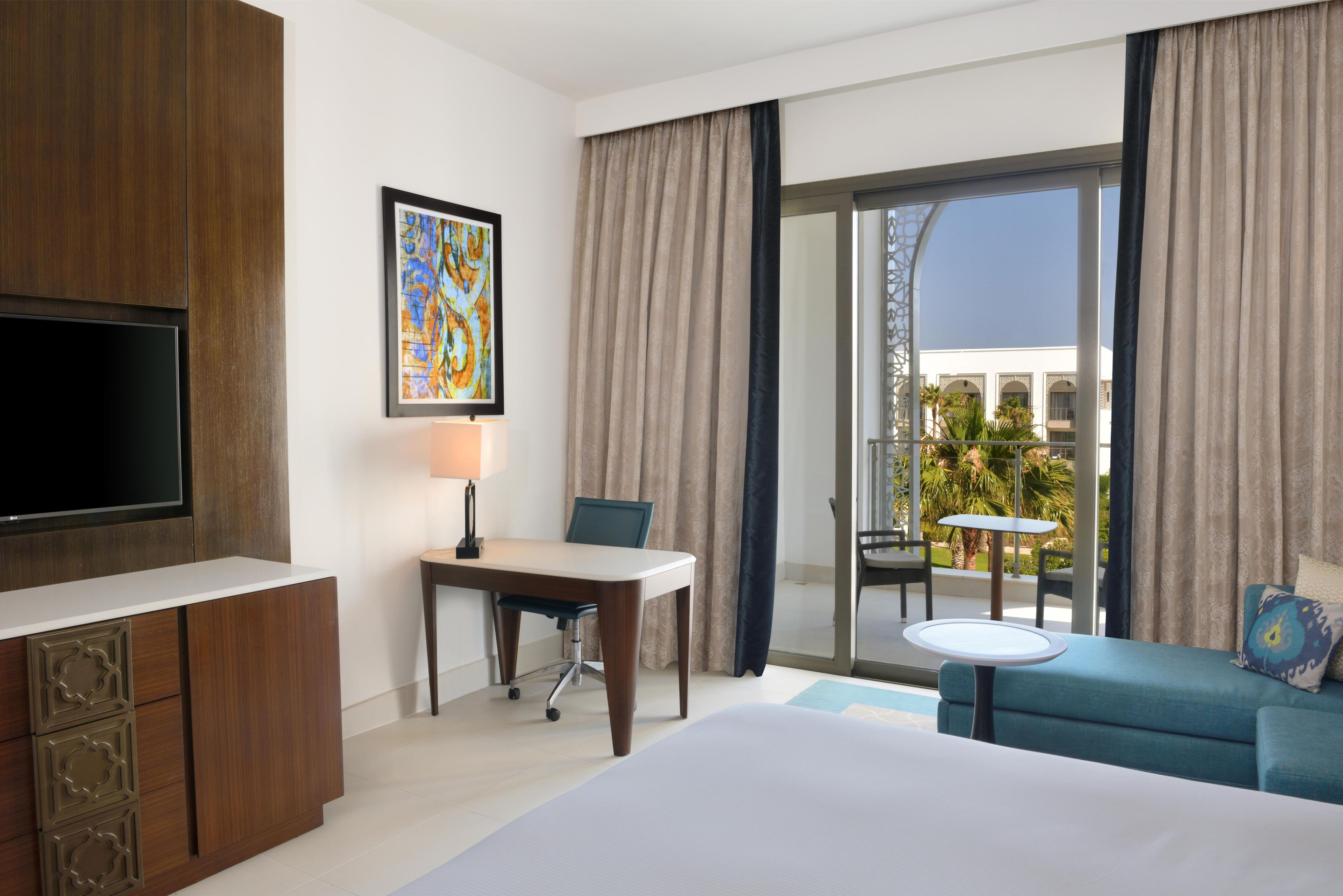Habitación de hotel con cama, escritorio, silla, televisor, balcón con mesa y sillas, y vista al exterior.