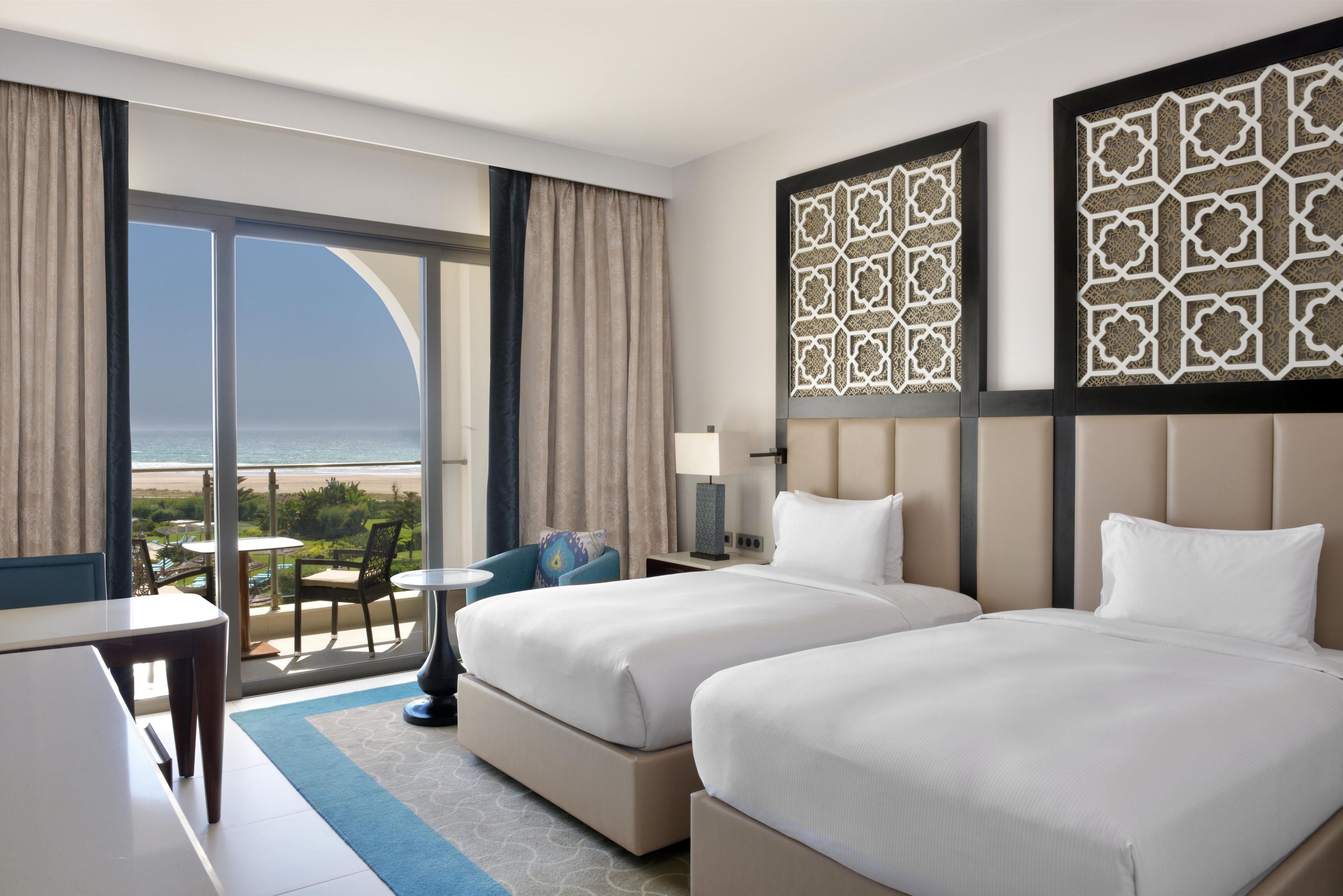A hotel room with two beds, a balcony with a view of the beach, and decorative wall panels.
