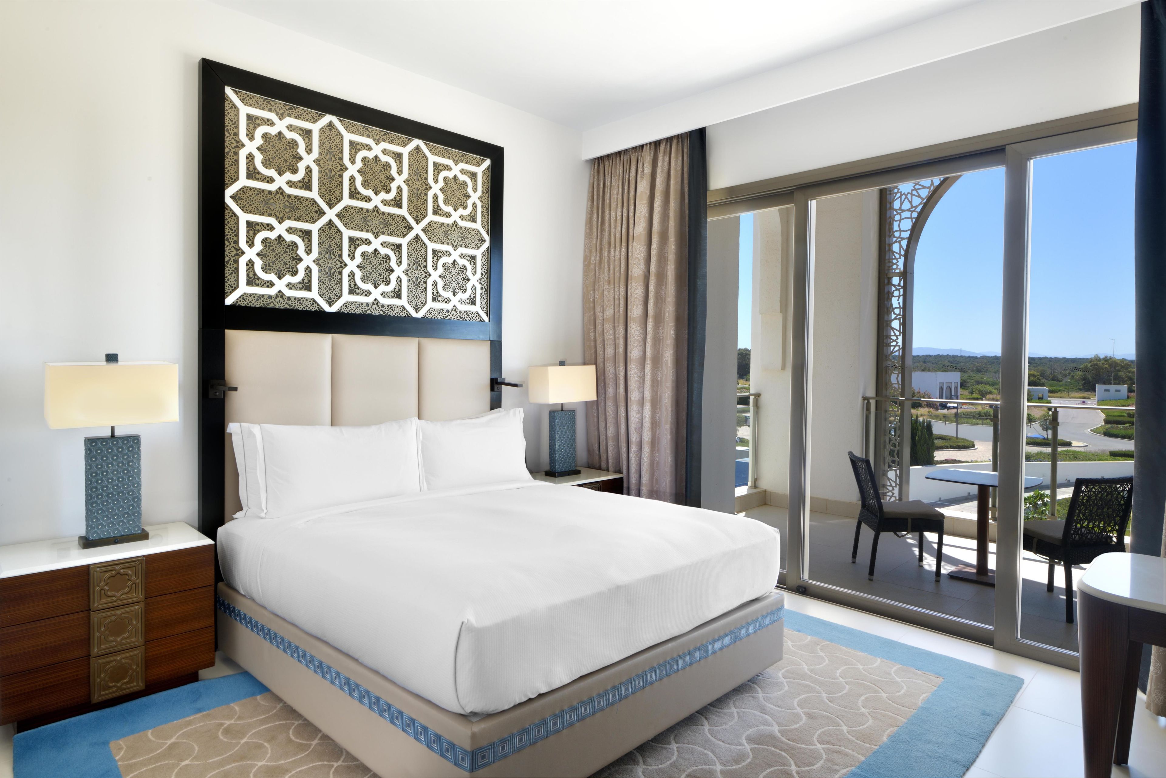 Habitación de hotel con una cama grande, dos mesitas de noche con lámparas, un panel decorativo en la pared, una puerta corredera de vidrio a un balcón con mobiliario exterior y vistas a la ciudad.
