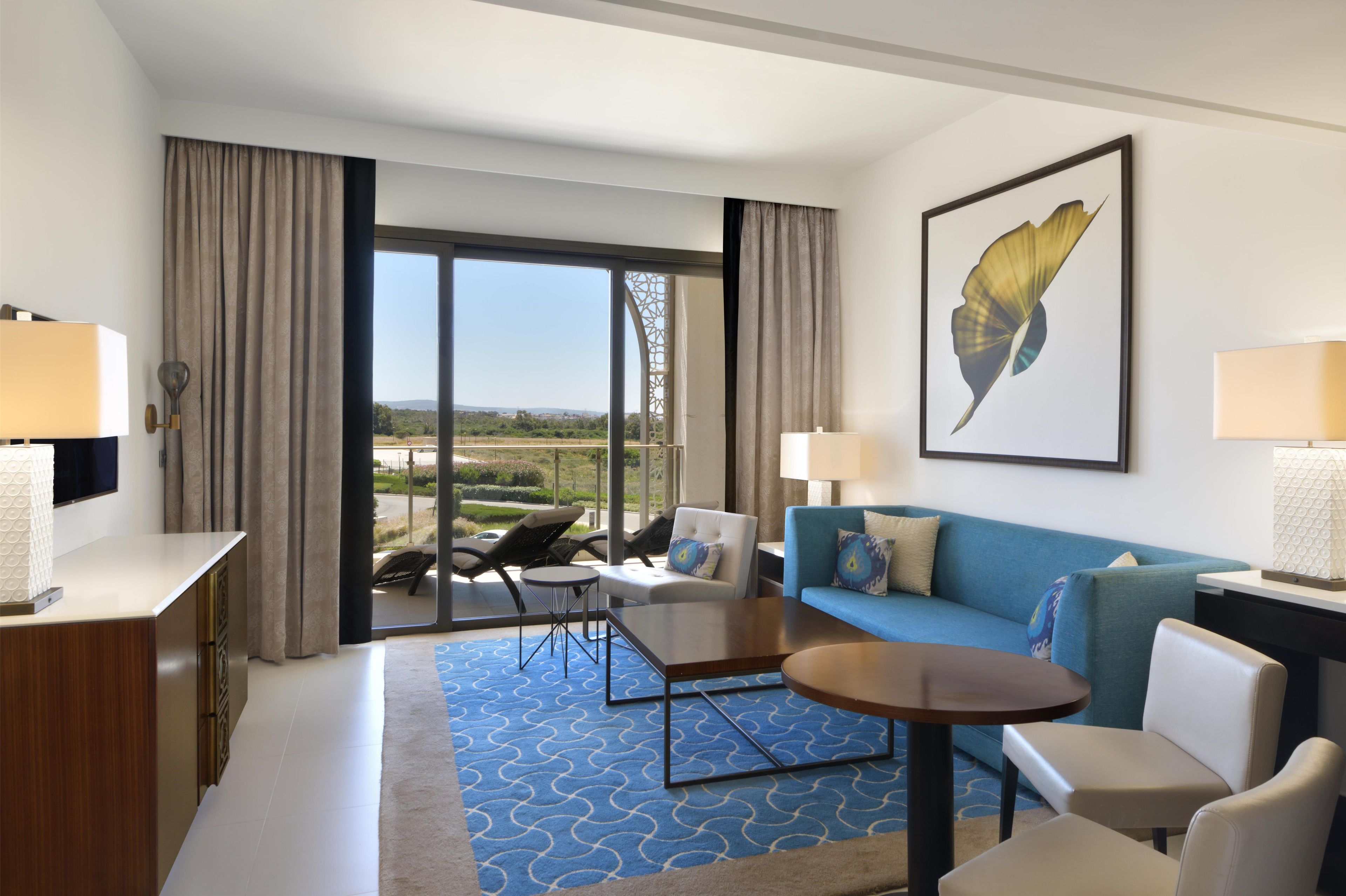 Una habitación de hotel con un ventanal grande, un sofá, una mesa de centro y un escritorio con lámpara.