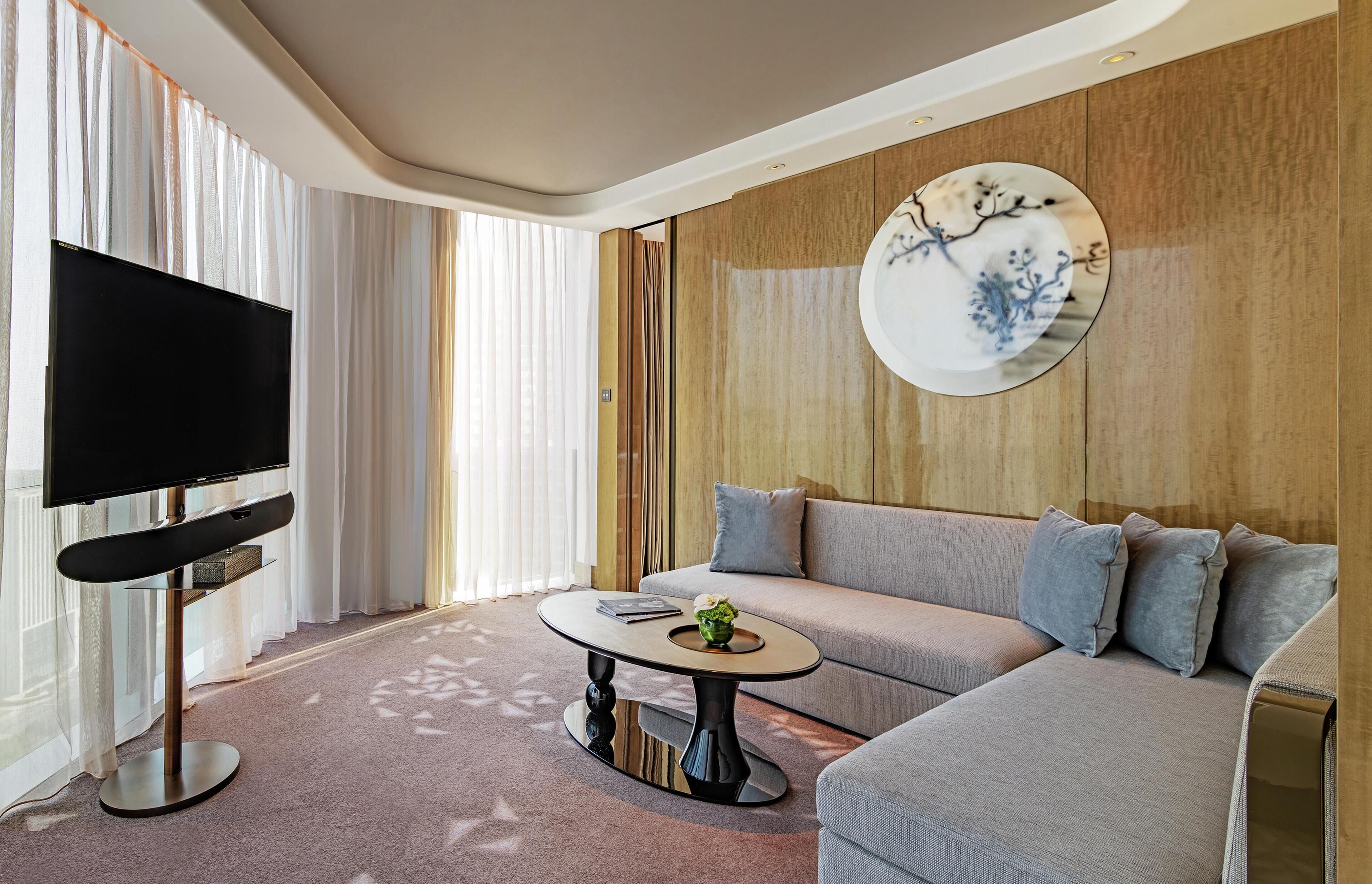 Un salon moderne avec un téléviseur à écran plat, une table basse ronde ornée d’un vase de fleurs, un canapé gris agrémenté de plusieurs coussins et une assiette décorative fixée au mur.