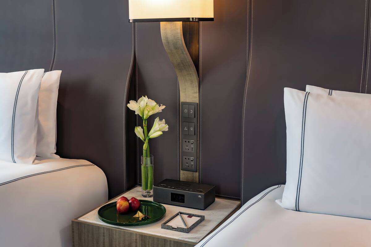 A hotel room with a bed, a bedside table with a lamp, a vase with flowers, and a plate with apples.