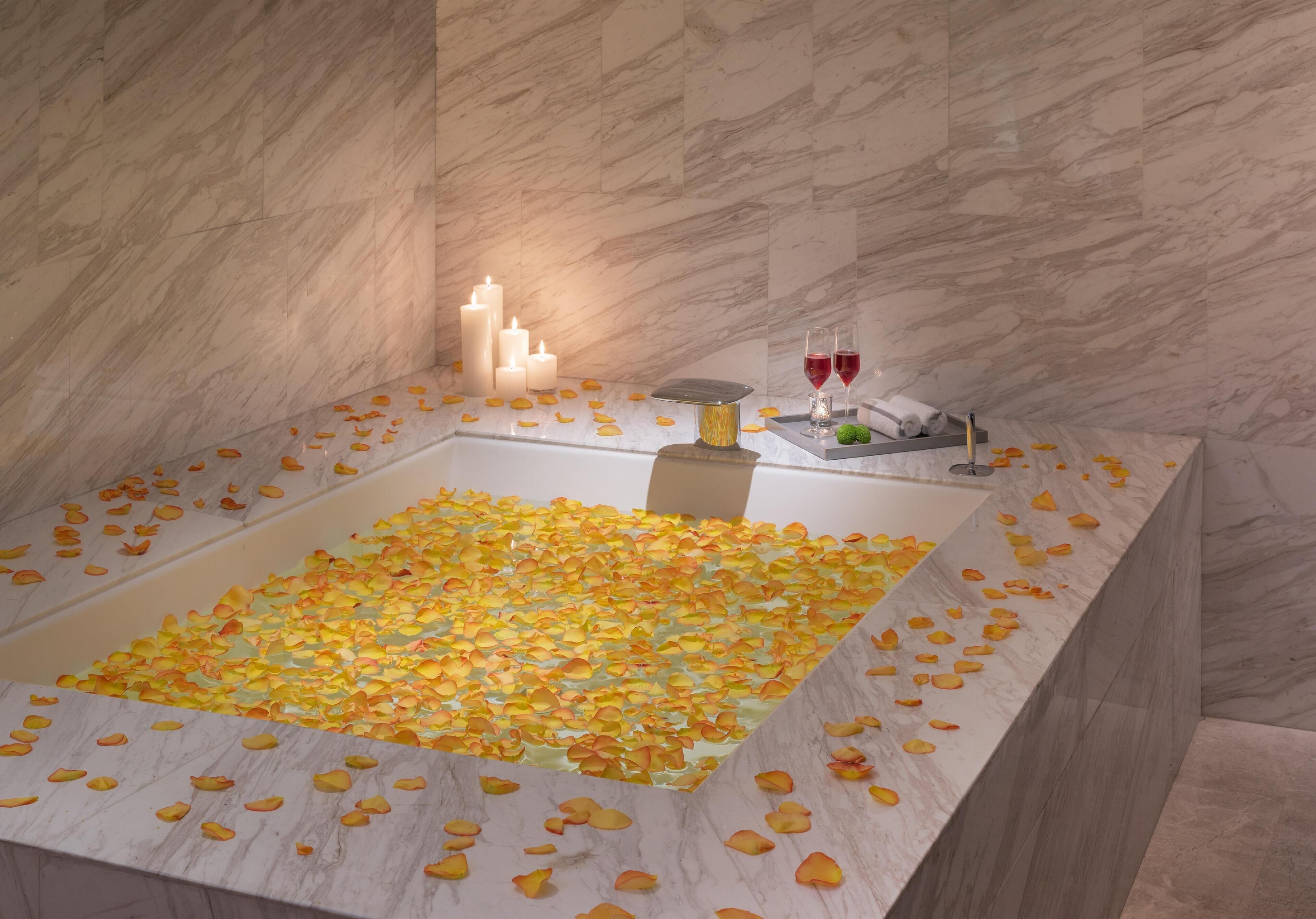 Una bañera llena de pétalos de rosa y velas, con una pared de mármol al fondo.