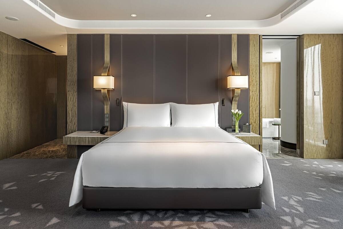 A modern hotel room with a large bed, two bedside lamps, a nightstand, and a small table with a vase of flowers.
