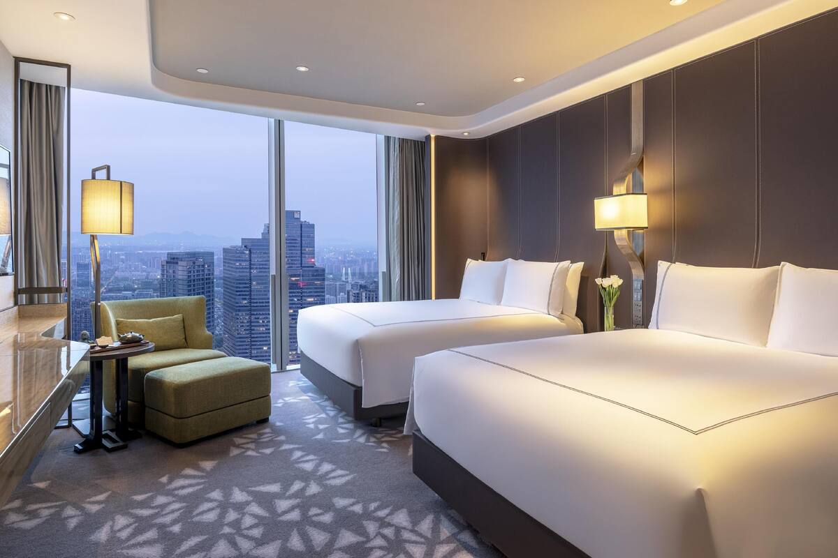 A hotel room with two beds, a sofa, a desk, and a large window offering a city view.