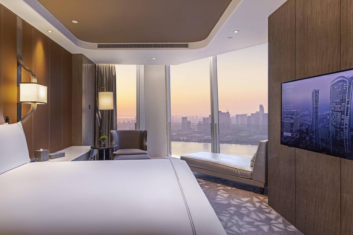 A hotel room with a large bed, a TV on the wall, a view of the cityscape, and a window with curtains.