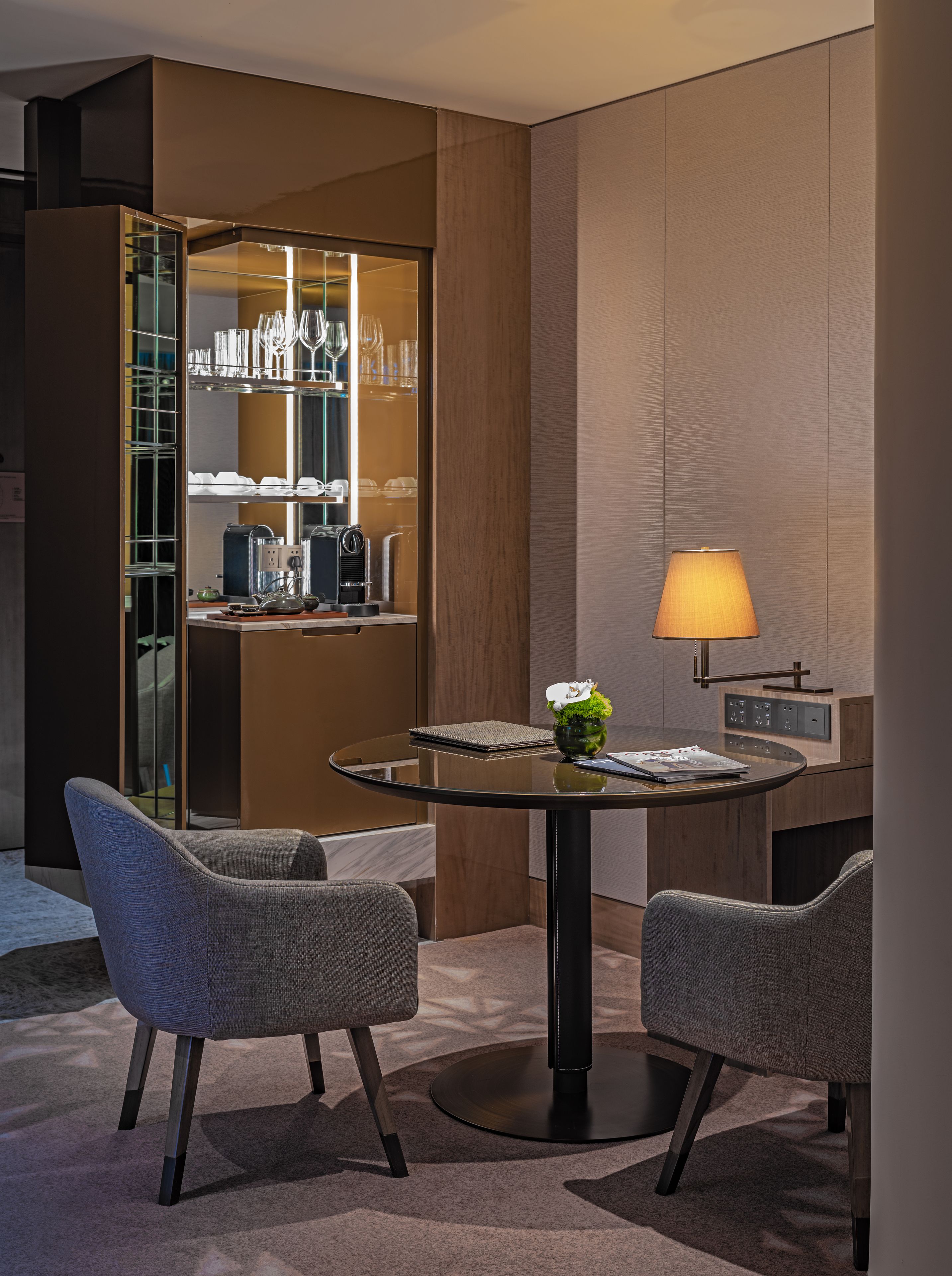 A modern hotel room with a round glass table, two grey upholstered chairs, a glass cabinet with kitchenware, a lamp, and a small plant.