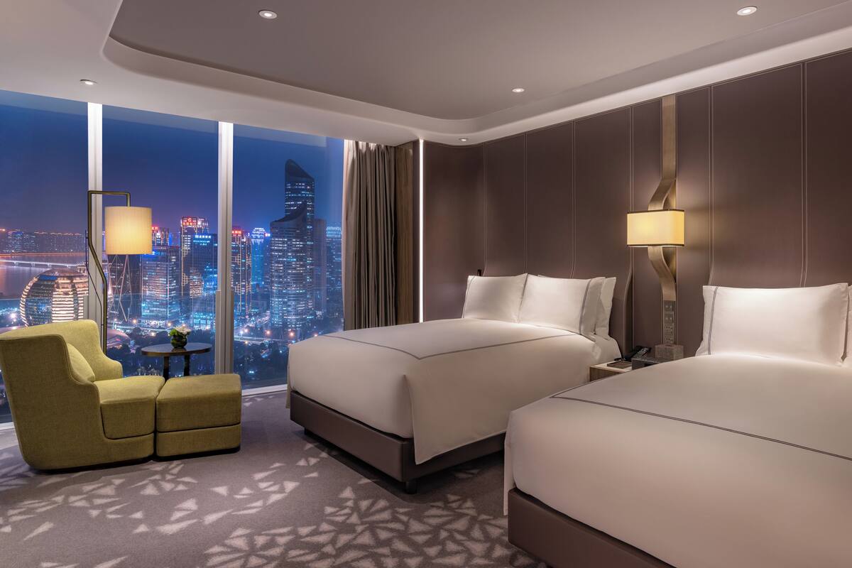 A hotel room with two beds, a sofa, a small table, and a view of a cityscape at night.