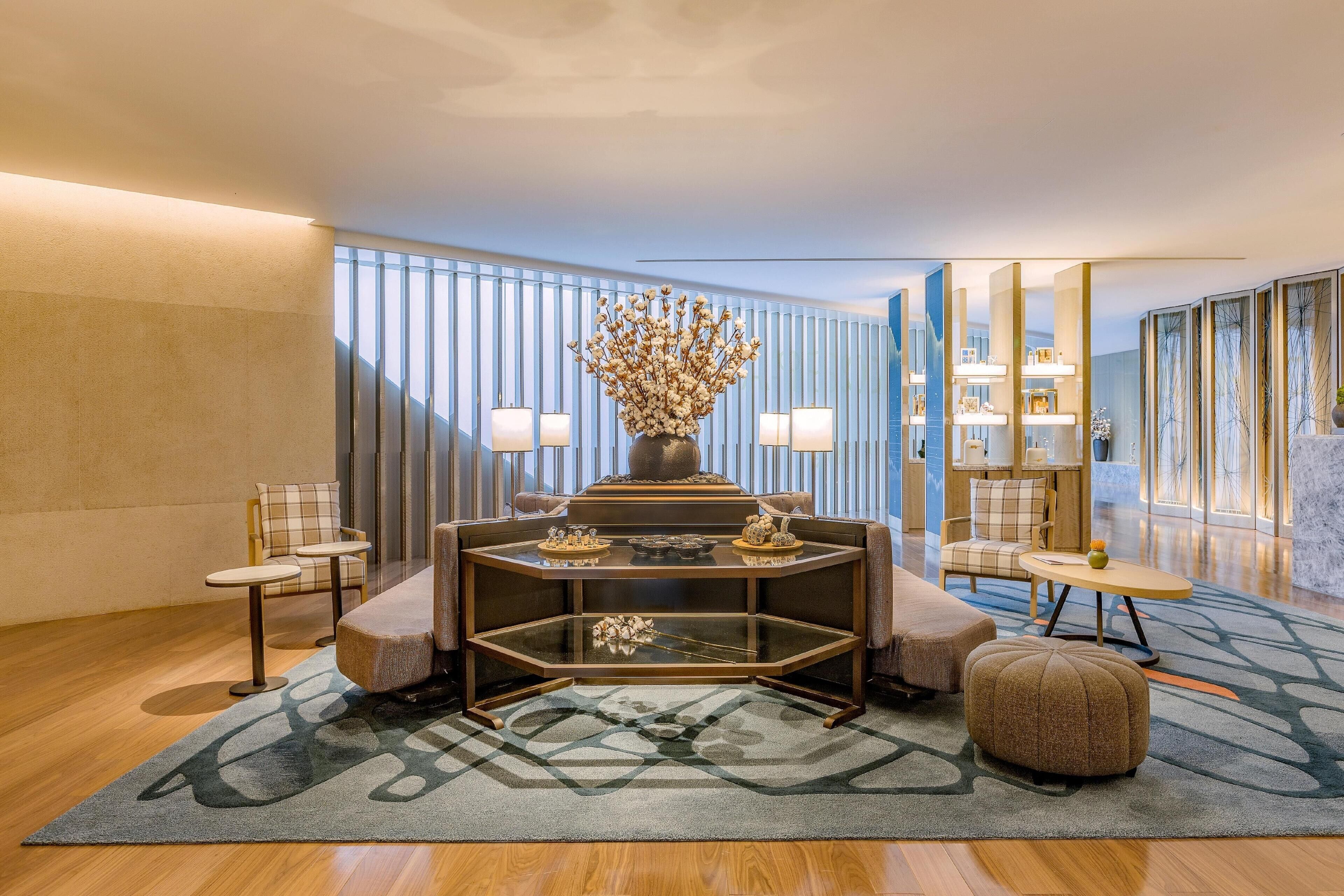 Un moderno vestíbulo de hotel con un área de estar central que incluye una mesa con cubierta de vidrio, dos sillones y un sofá. Un jarrón decorativo con flores secas está sobre la mesa. La habitación tiene un gran ventanal con persianas verticales, una alfombra estampada y piso de madera.