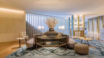 A modern hotel lobby with a central seating area featuring a glass-top table, two armchairs, and a sofa. A decorative vase with dried flowers is on the table. The room has a large window with vertical blinds, a patterned rug, and a wooden floor.