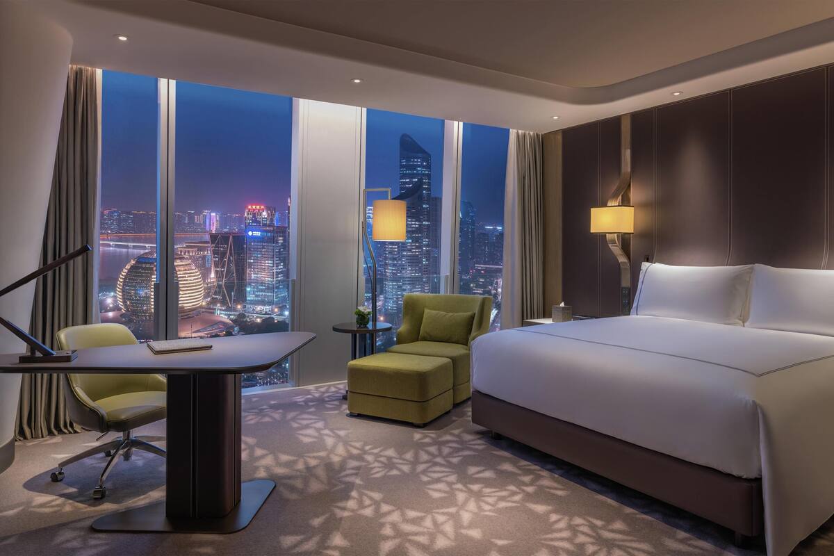 A hotel room with a large bed, a desk with a chair, a small table, and a view of a cityscape at night.