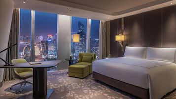 Habitación de hotel con una cama grande, un escritorio con una silla, una mesita y vistas a un paisaje urbano nocturno.