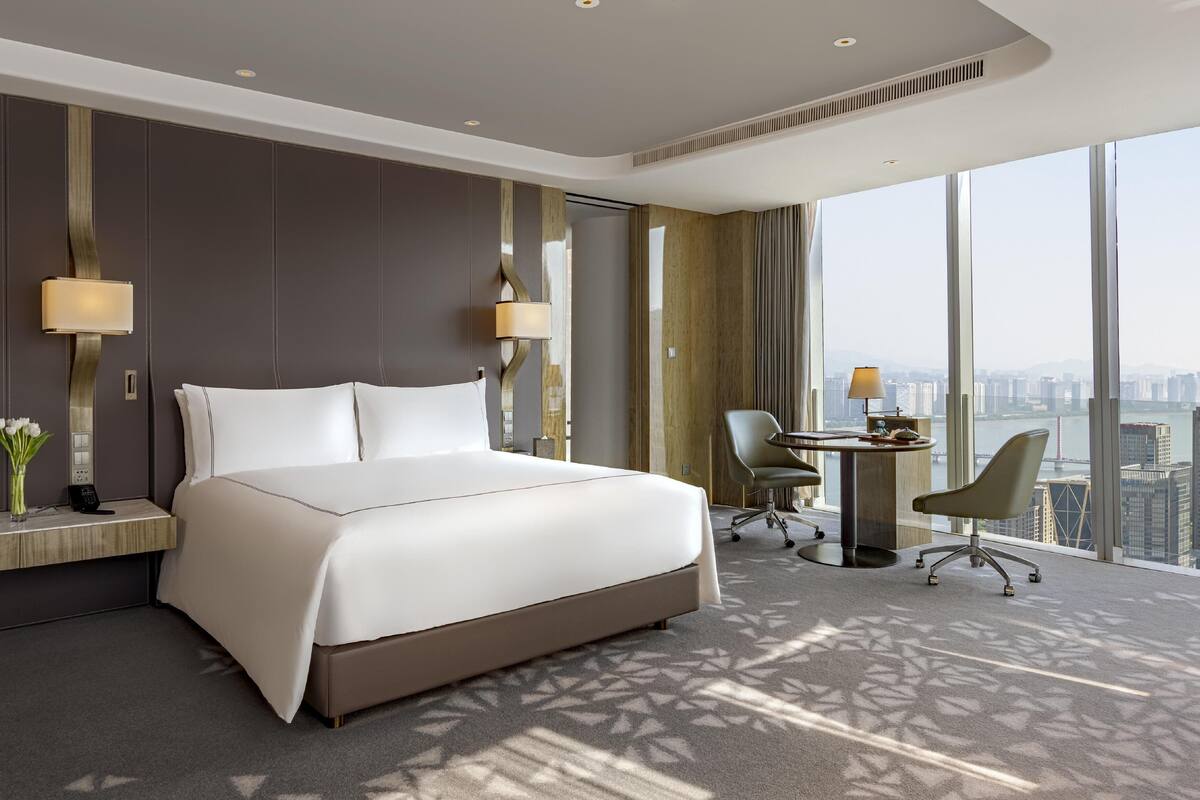 A modern hotel room with a large bed, a desk with a chair, and a view of the cityscape through large windows.