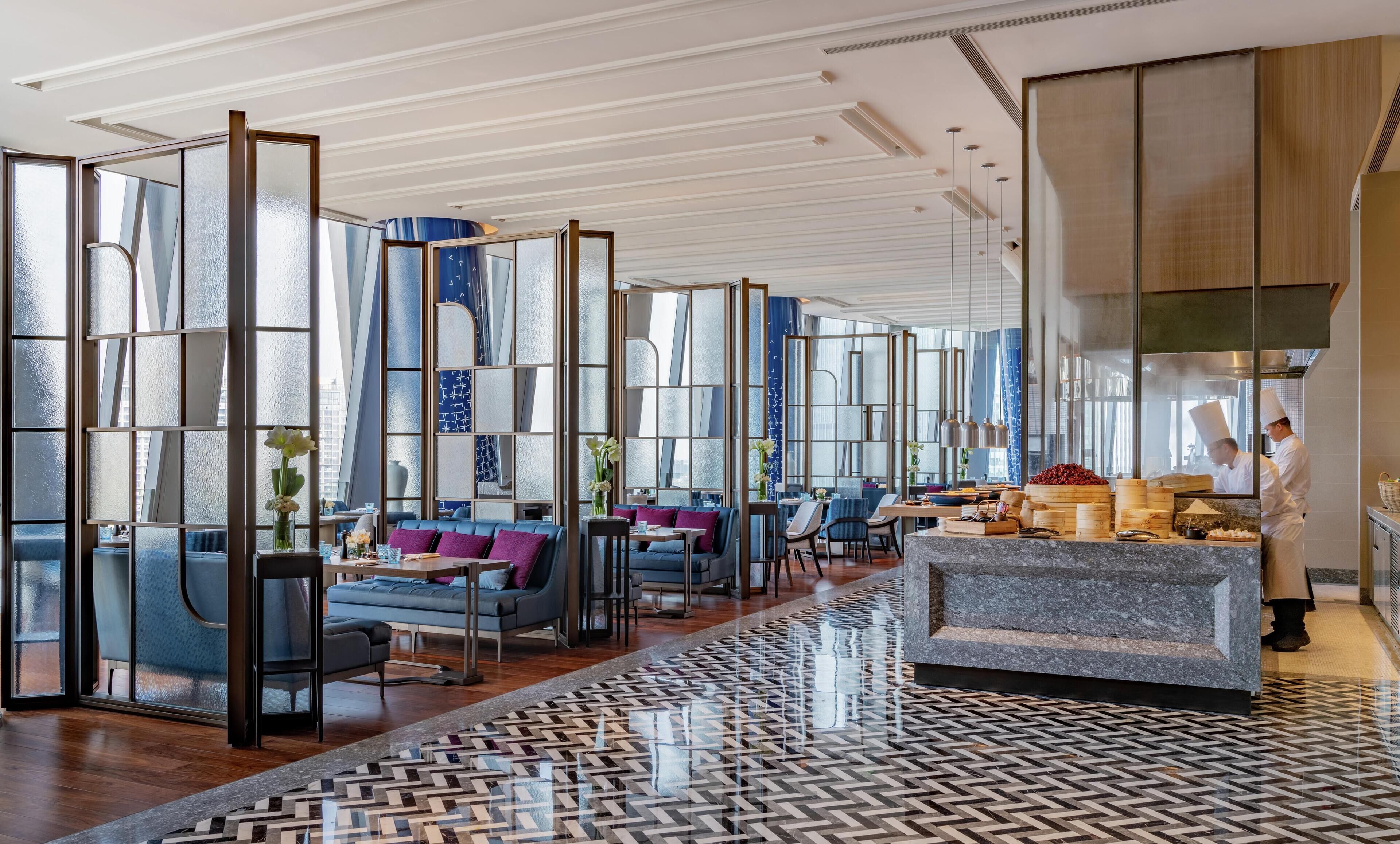 A modern hotel lobby with seating areas, a kitchen visible through a glass partition, and a patterned floor.