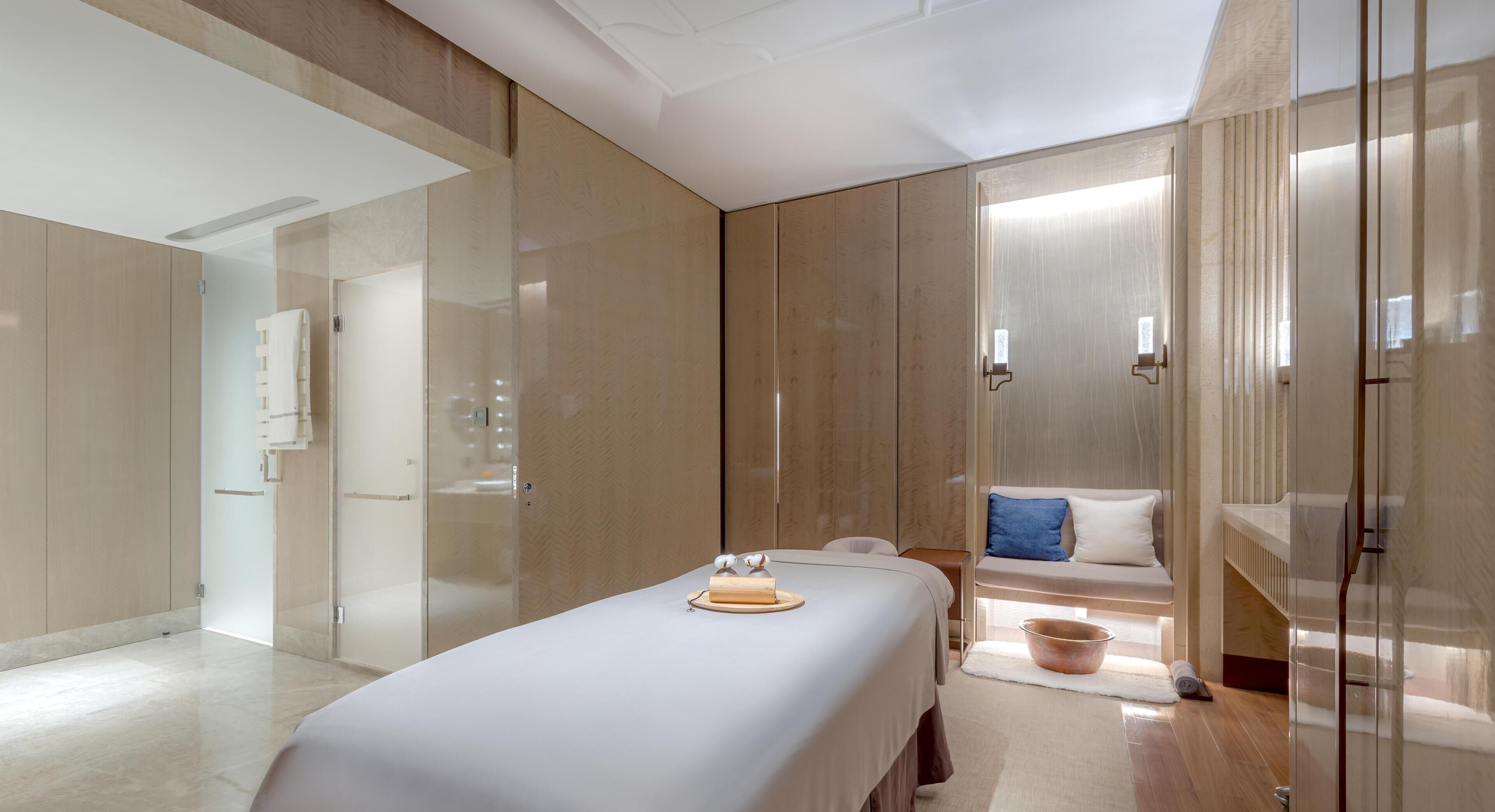 Une chambre d’hôtel moderne avec un grand lit, un petit coin salon avec un repose-pieds, et une cloison en verre.
