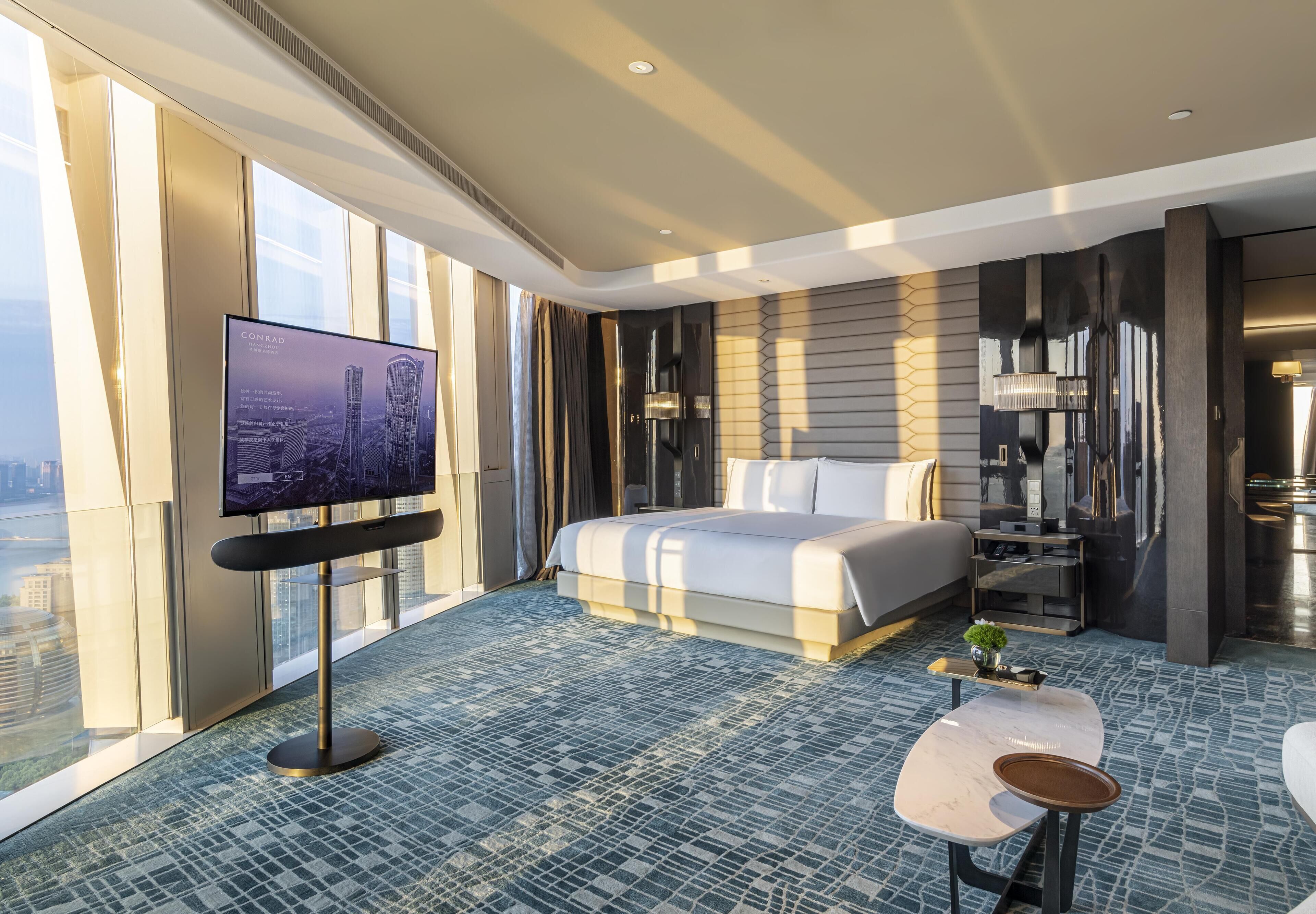A modern hotel room with a large bed, a TV on a stand, a small table, and a view of the city through large windows.