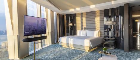 A modern hotel room with a large bed, a TV on a stand, a small table, and a view of the city through large windows.