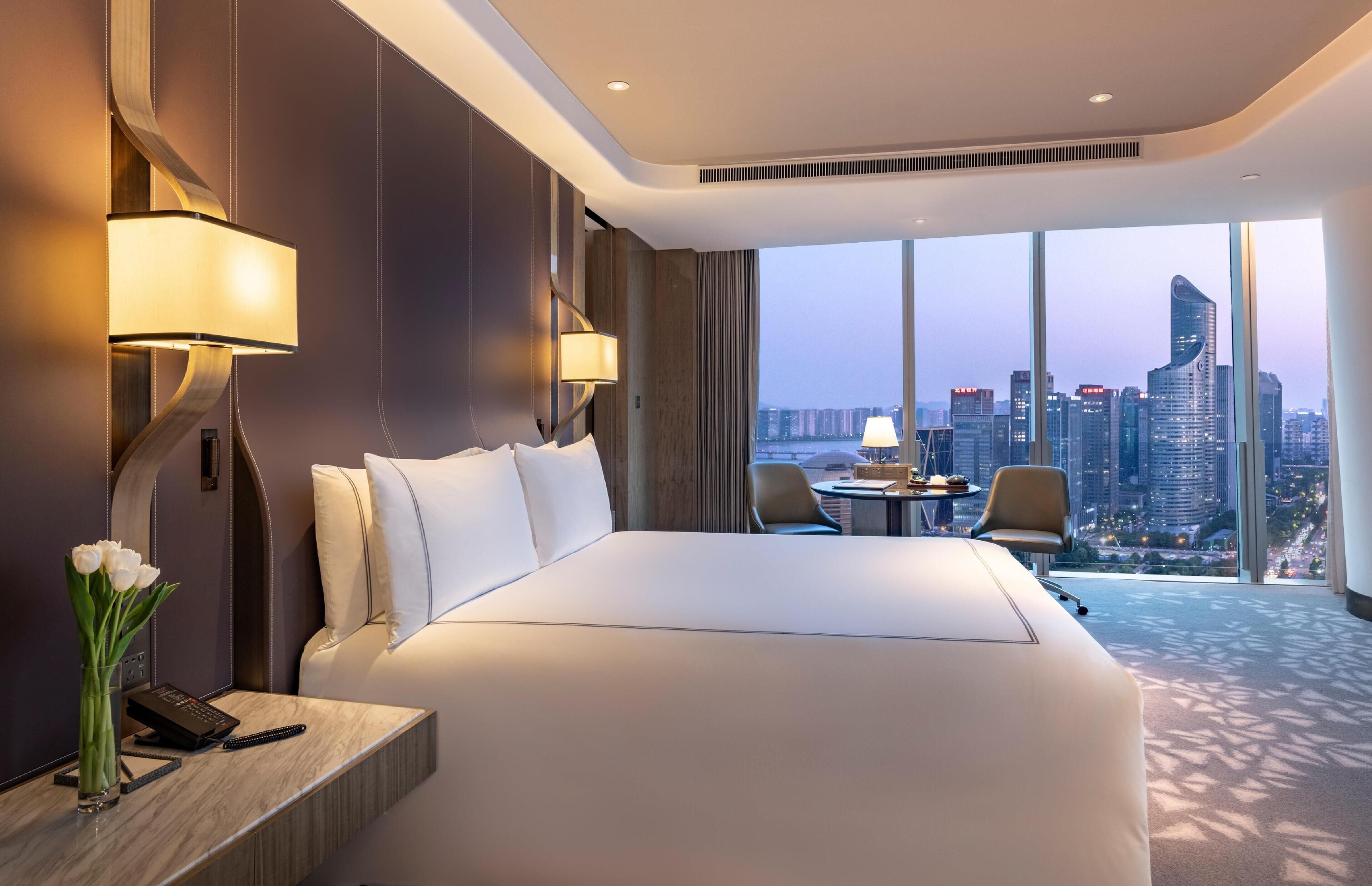 A hotel room with a large bed, a desk with a lamp, a vase of flowers, and a view of a city skyline through large windows.