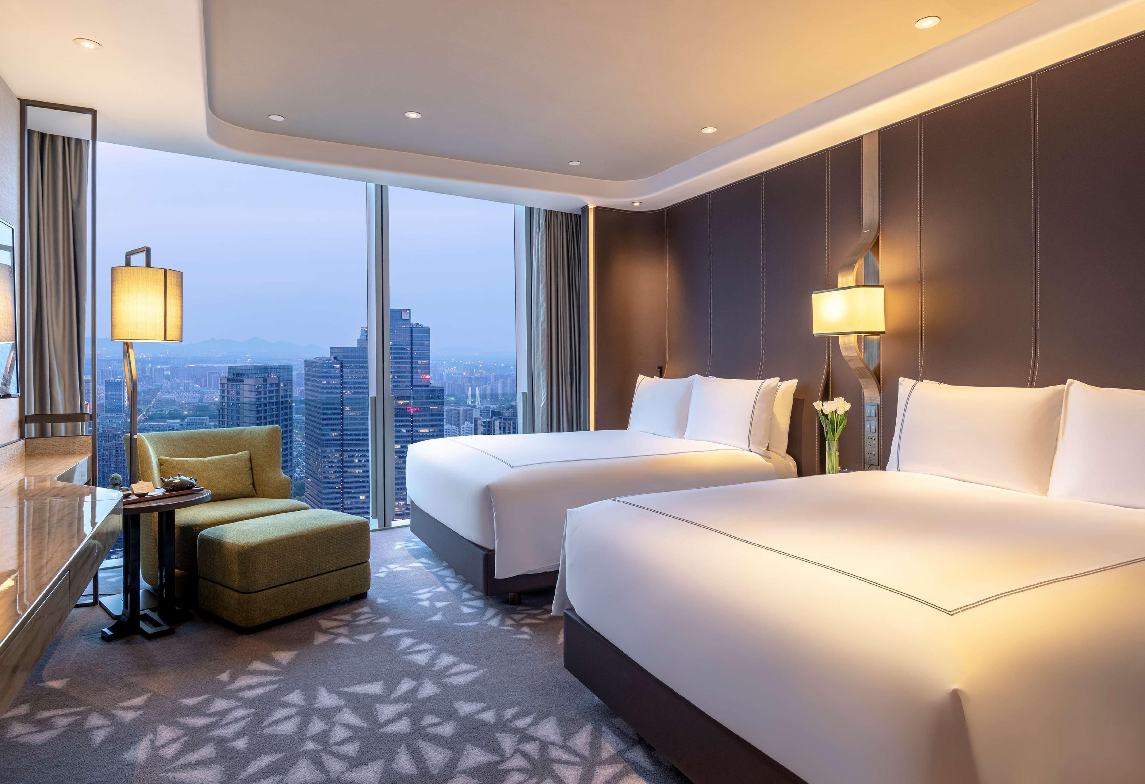 A hotel room with two beds, a sofa, a desk, and a view of the cityscape.