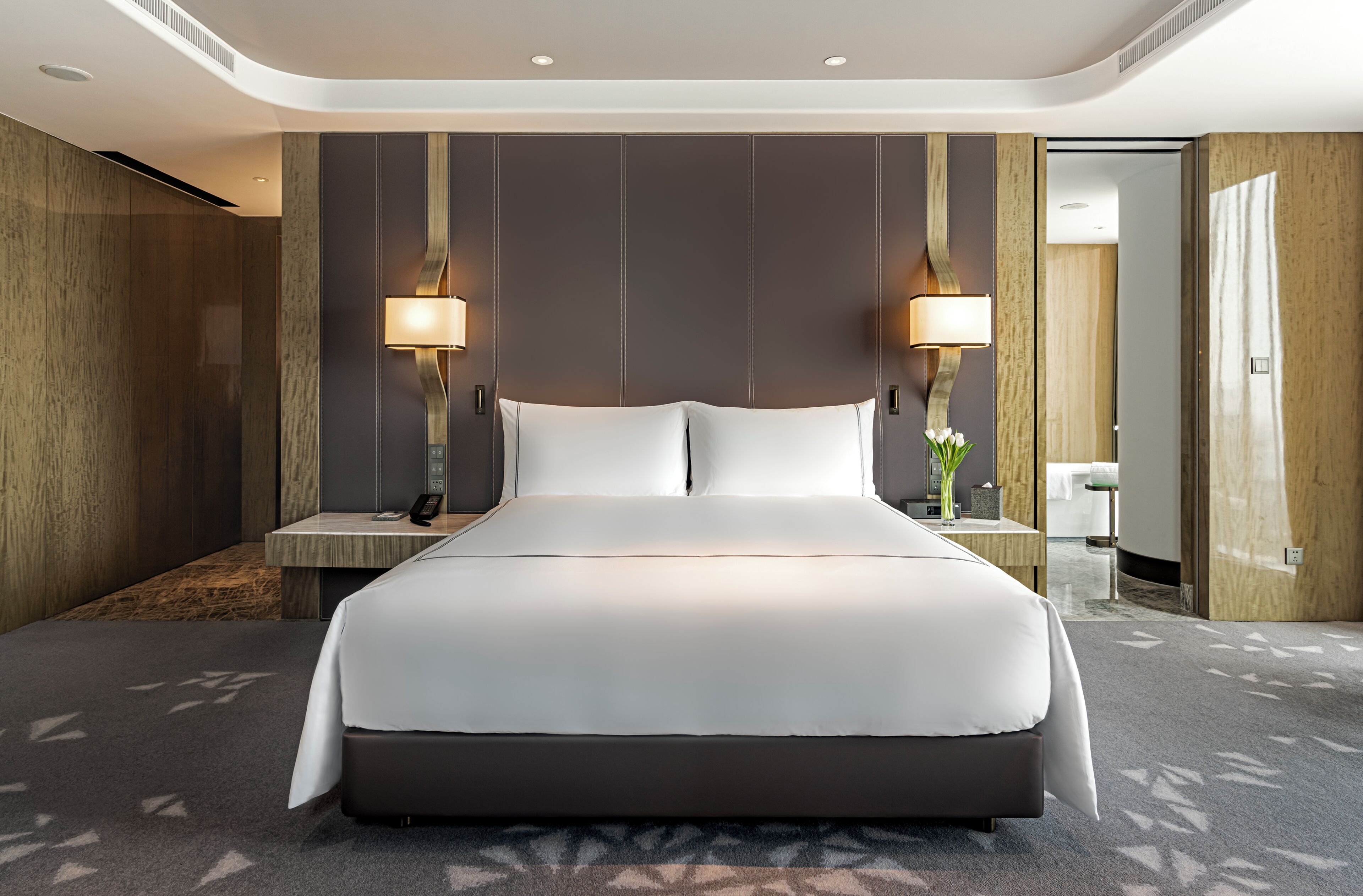 Una habitación de hotel moderna con una cama grande, dos lámparas de noche, una mesita de noche y un baño visible a través de una puerta abierta.