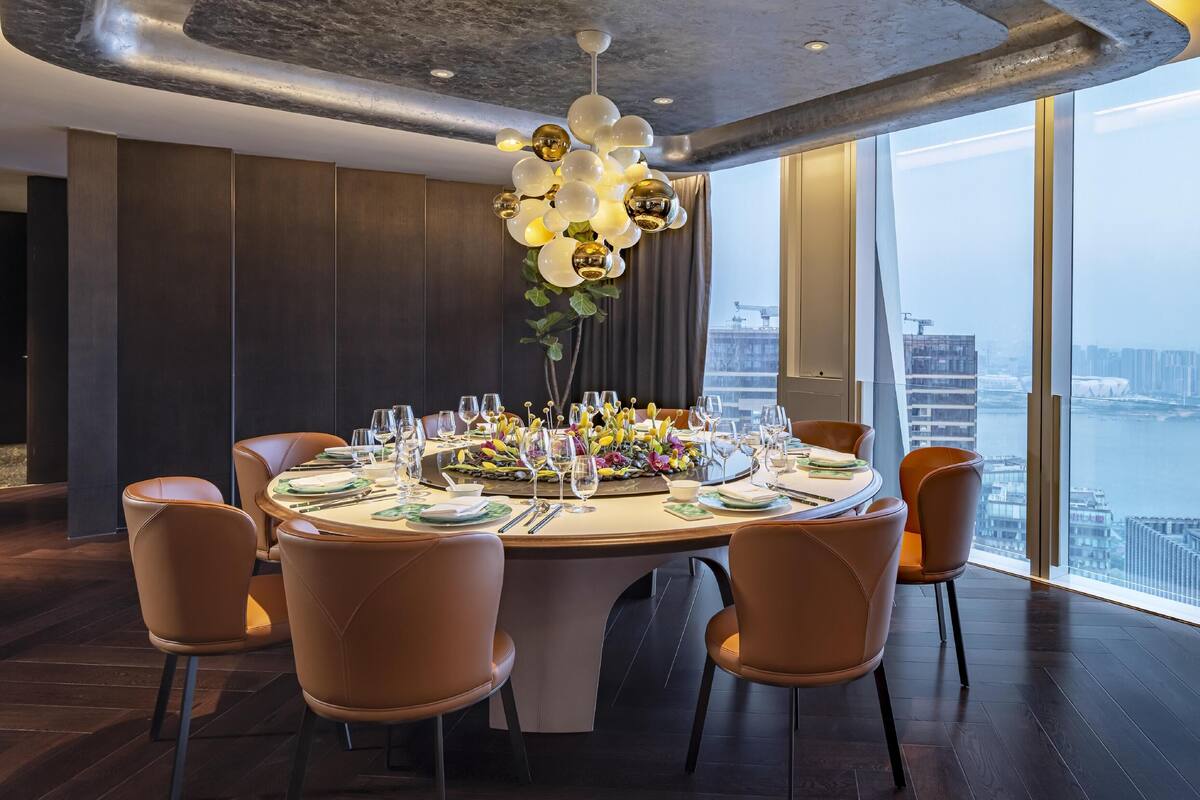 A dining area with a round table set for six, surrounded by six brown chairs. The table is decorated with a floral centerpiece, plates, glasses, and cutlery. The room has a modern design with dark wood flooring, a high ceiling with exposed beams, and large windows offering a view of a cityscape and a body of water.