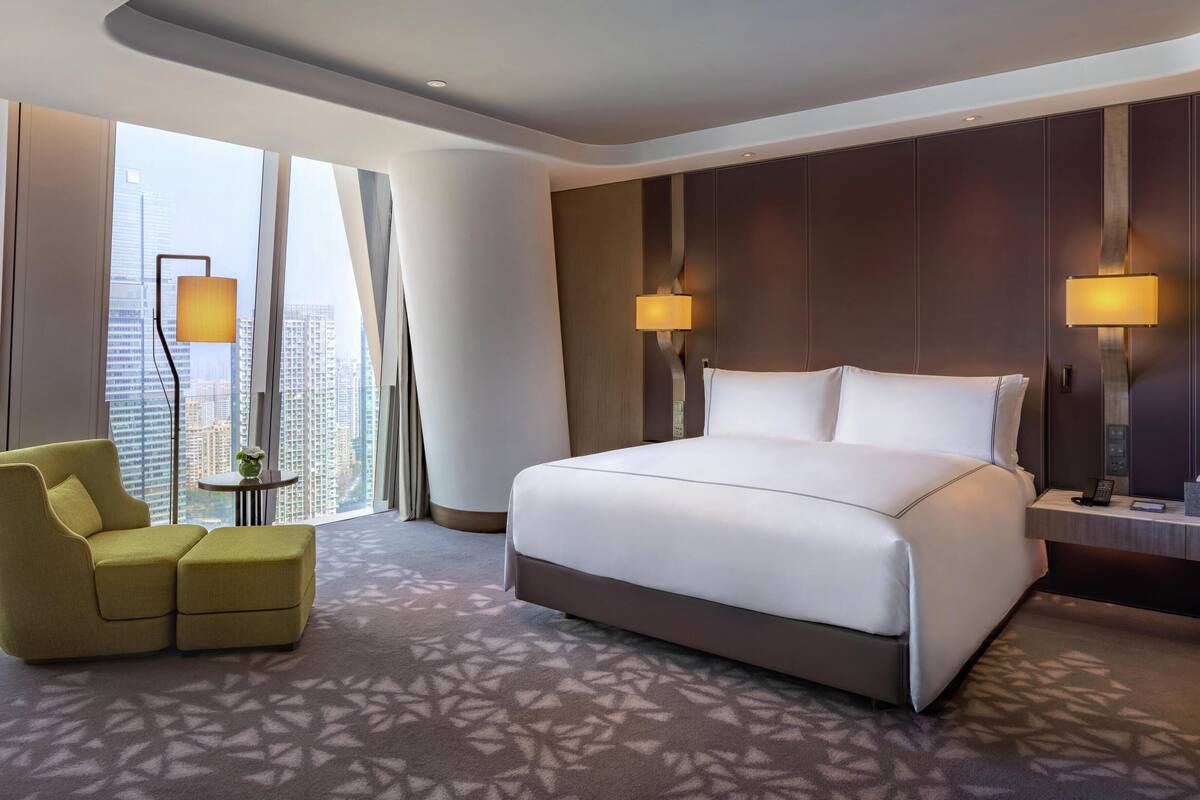 A modern hotel room with a large bed, a seating area, and a view of the cityscape.