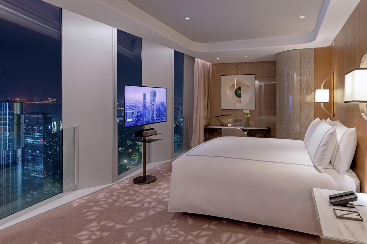 A modern hotel room with a large bed, a desk, a chair, a TV, and a city view.