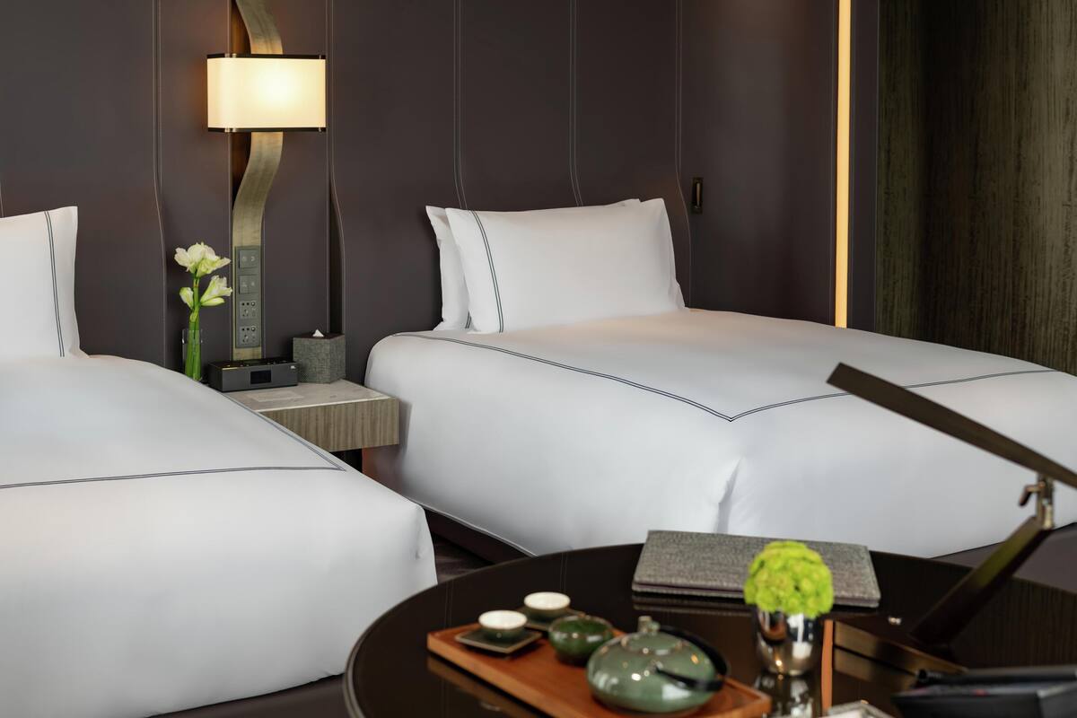 A hotel room with two beds, a bedside table with a lamp, a small vase with flowers, and a tray with teapots and cups.