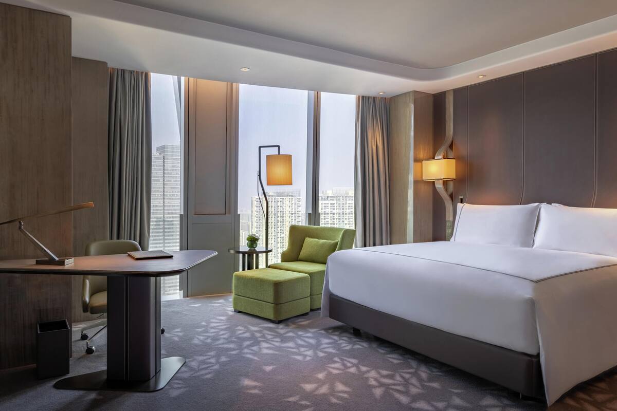 A modern hotel room with a large bed, a desk, a green armchair, and a view of the cityscape through large windows.