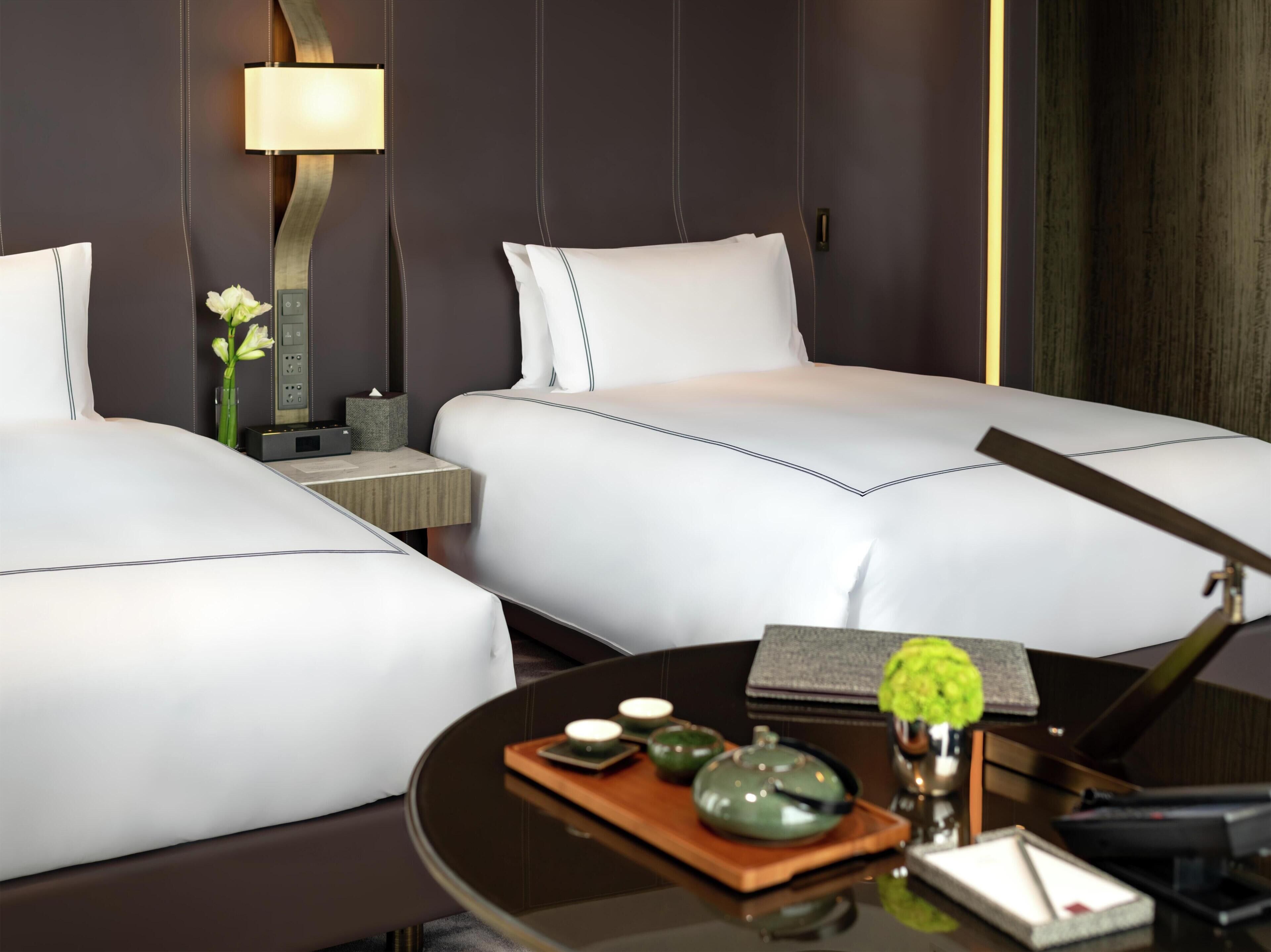 A hotel room with two beds, a bedside table with a lamp, a vase with flowers, and a tray with teapots and cups.