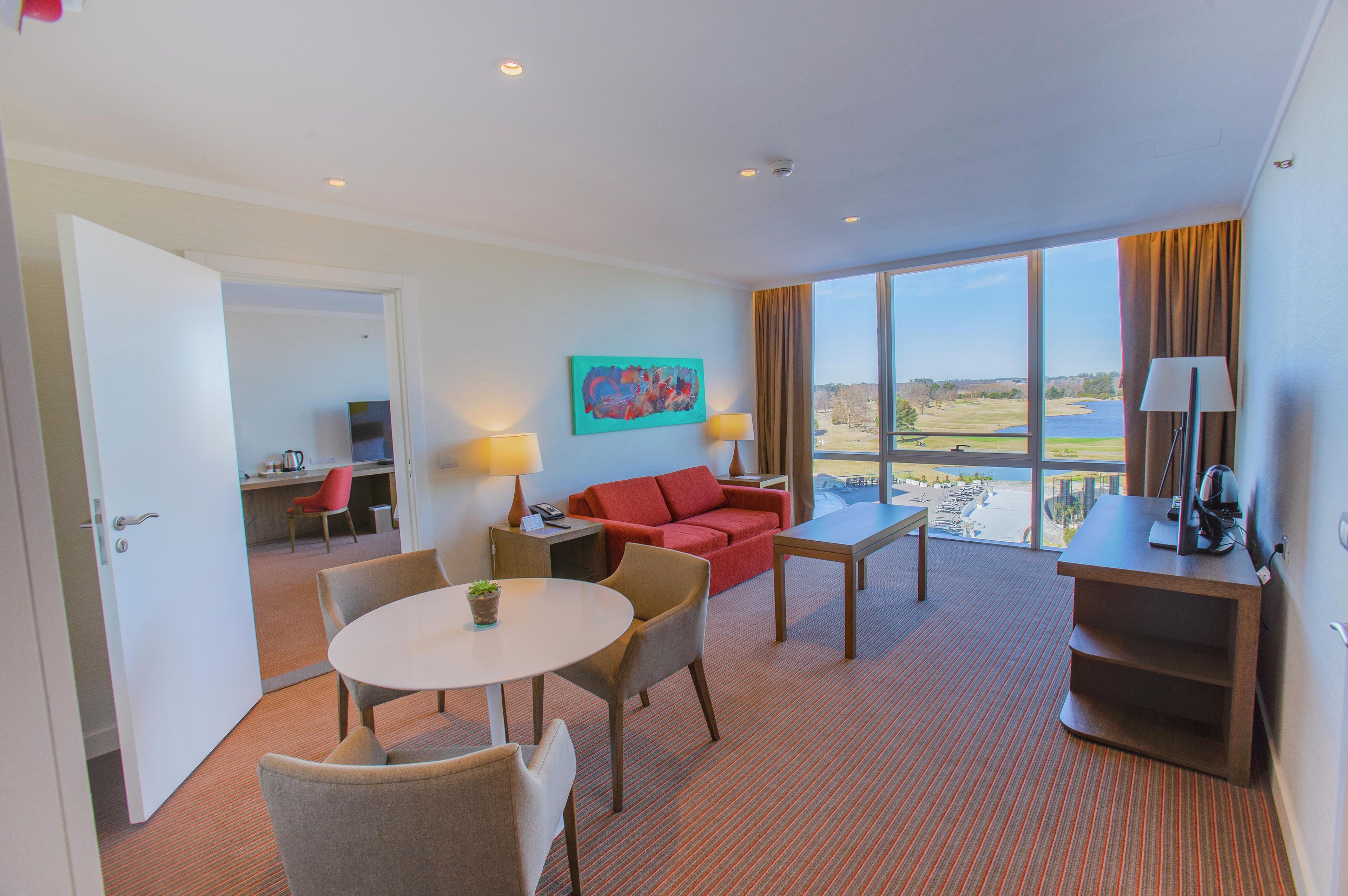 Habitación de hotel con zona de comedor, un sofá rojo, un cuadro y vista a un campo de golf.