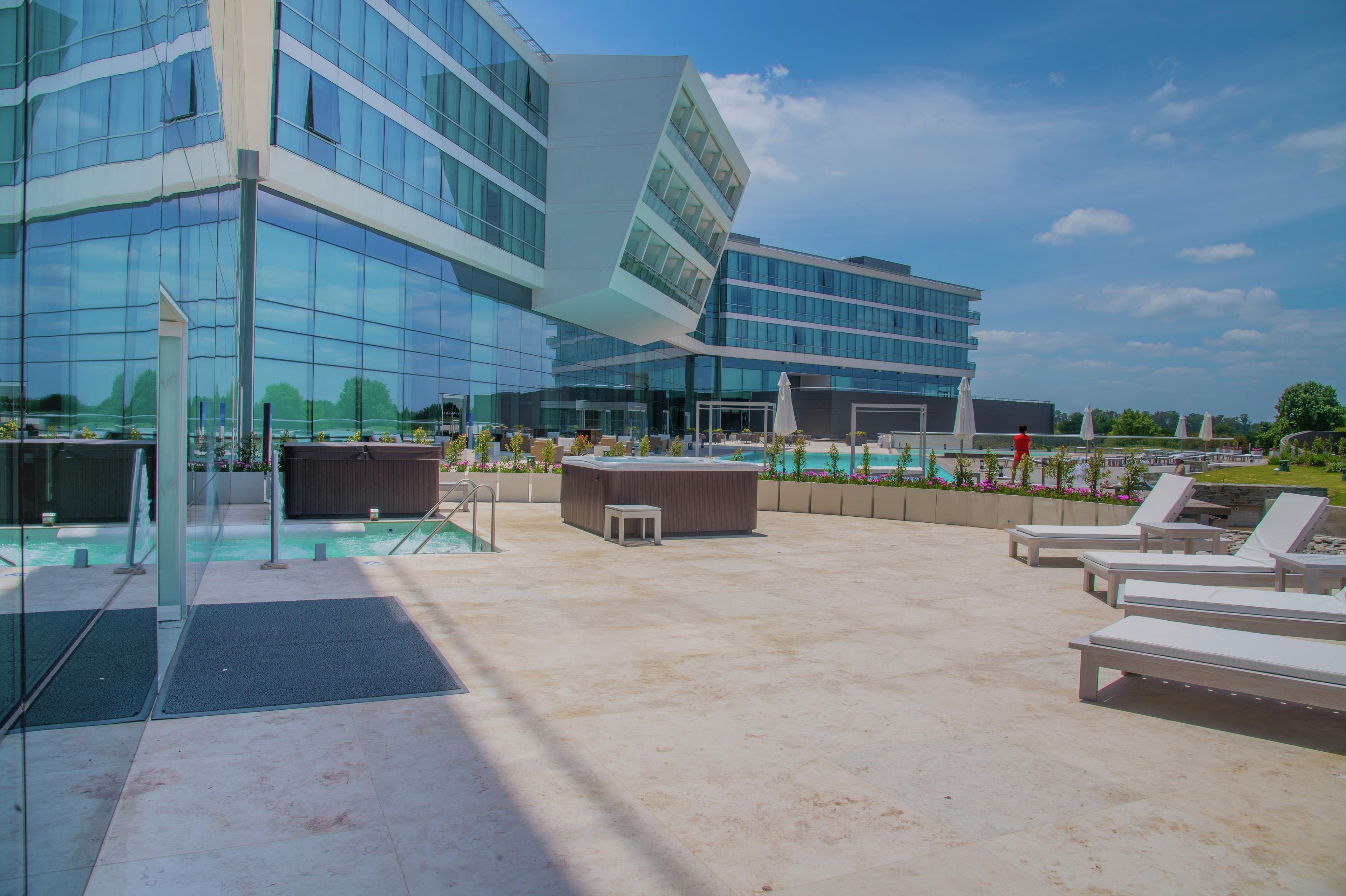 A rooftop pool area with lounge chairs, a hot tub, and a modern building with glass windows.