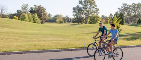 Dos personas montando en bicicleta sobre un camino pavimentado, con un área verde y árboles al fondo.