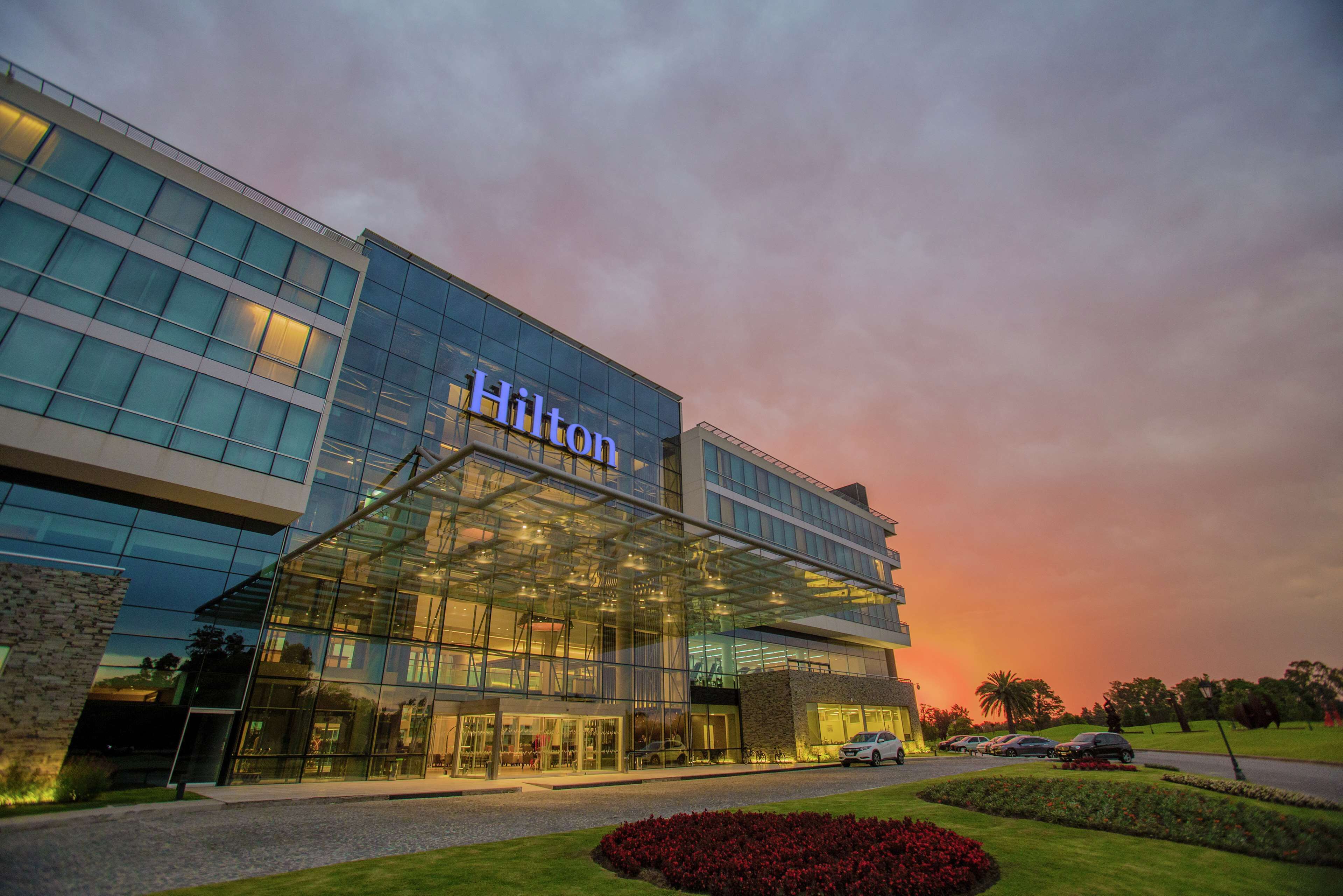 A modern Hilton hotel with a glass facade, well-lit entrance, and landscaped grounds.