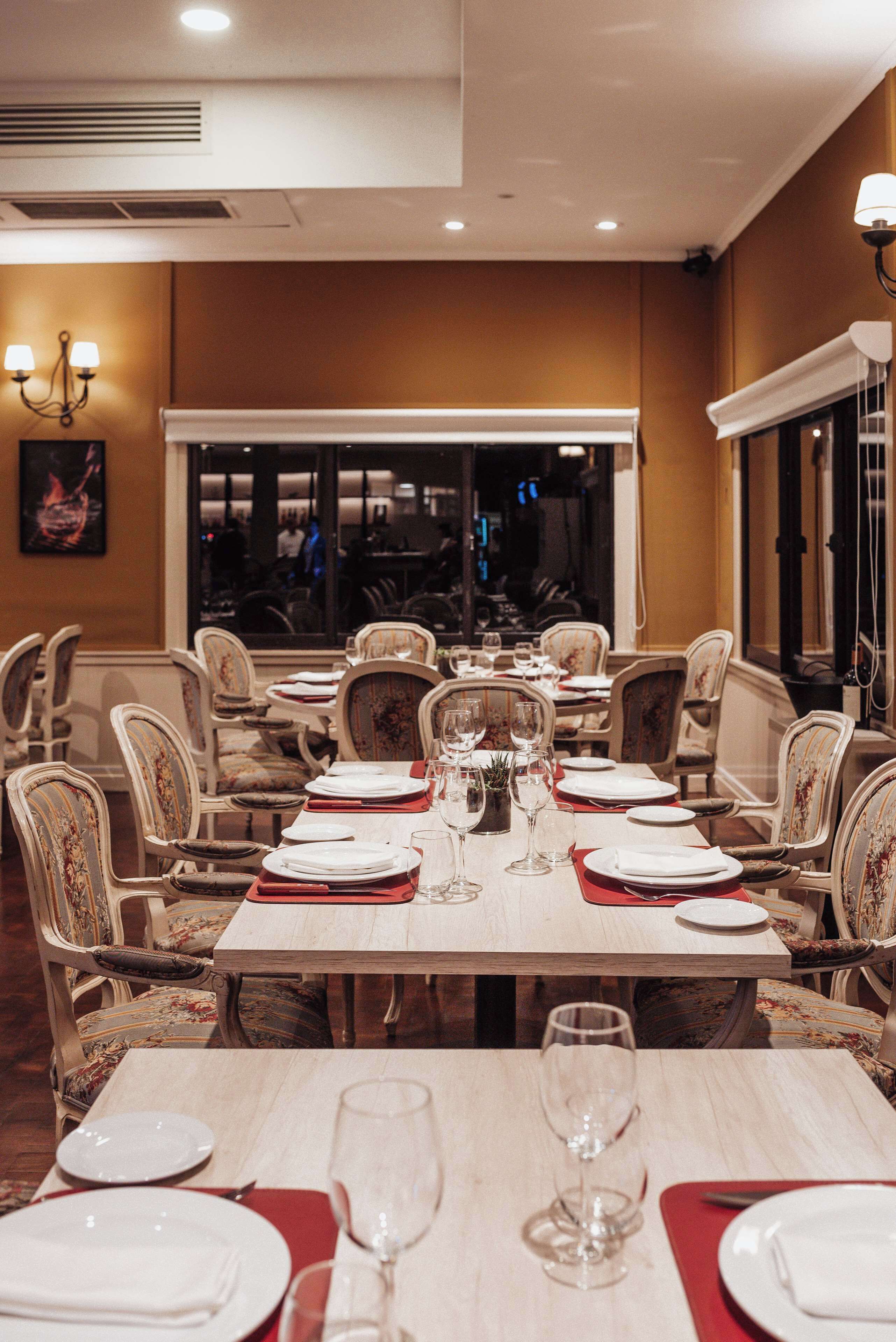 A restaurant with a long table set for dining, chairs with floral upholstery, and a view of the dining area through a large window.