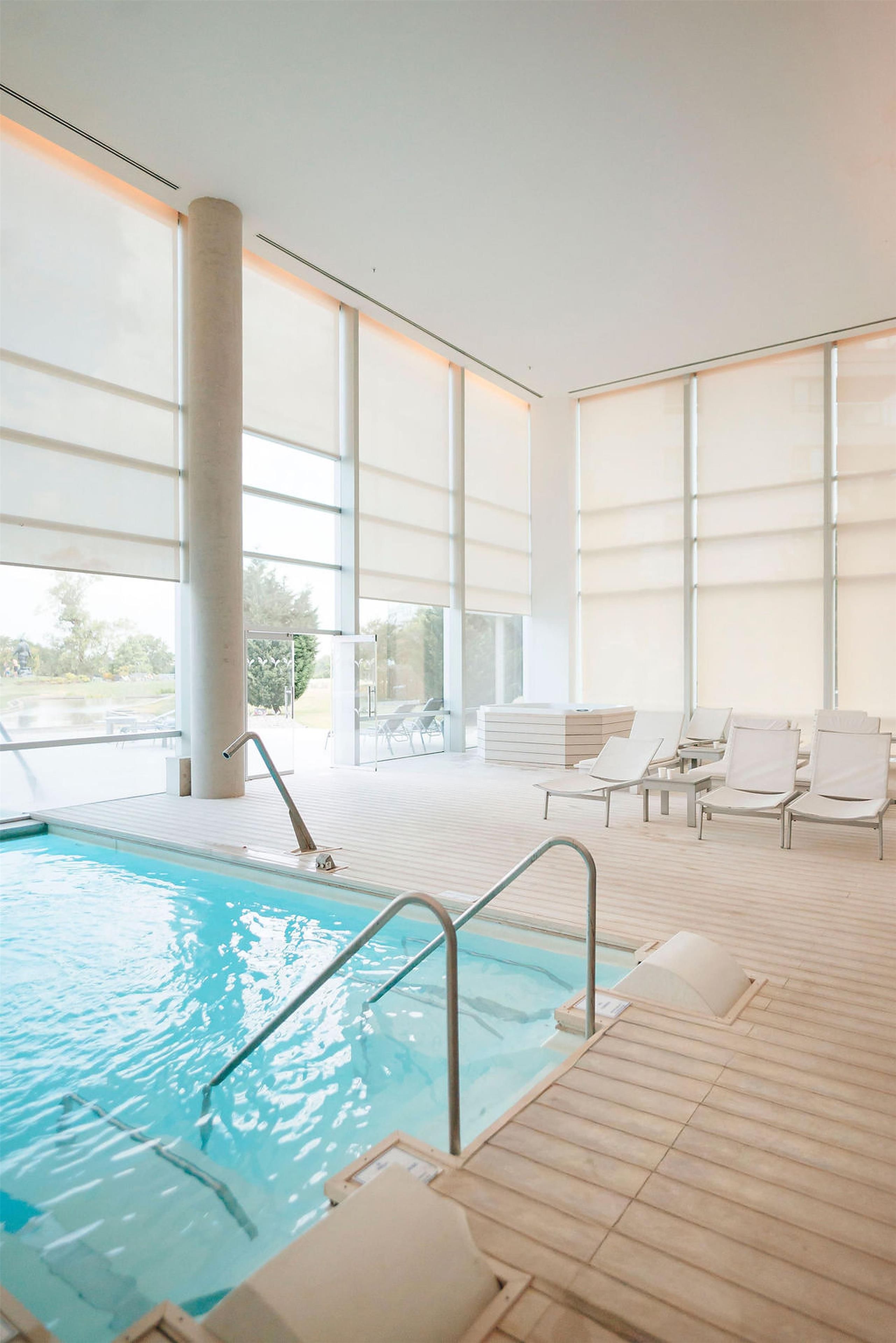 Une piscine intérieure avec des parois en verre transparent, des chaises longues et une terrasse en bois.