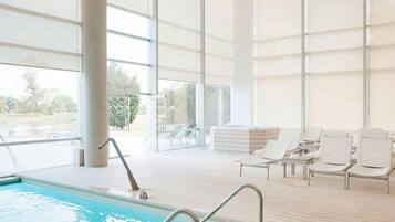 Une piscine intérieure avec des parois en verre transparent, des chaises longues et une terrasse en bois.