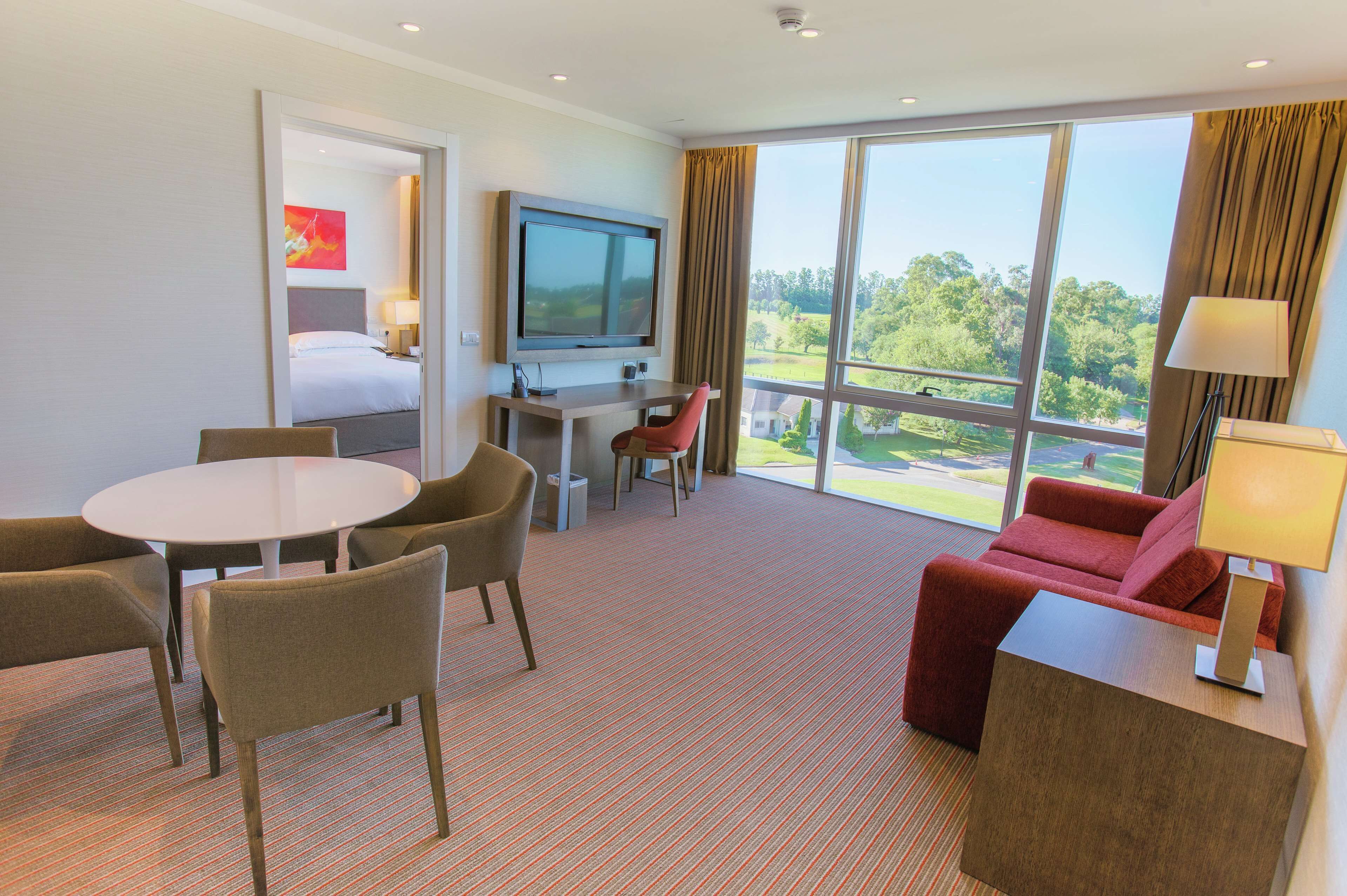 A hotel room with a bed, a TV, a desk, a red sofa, and a large window with a view of greenery.