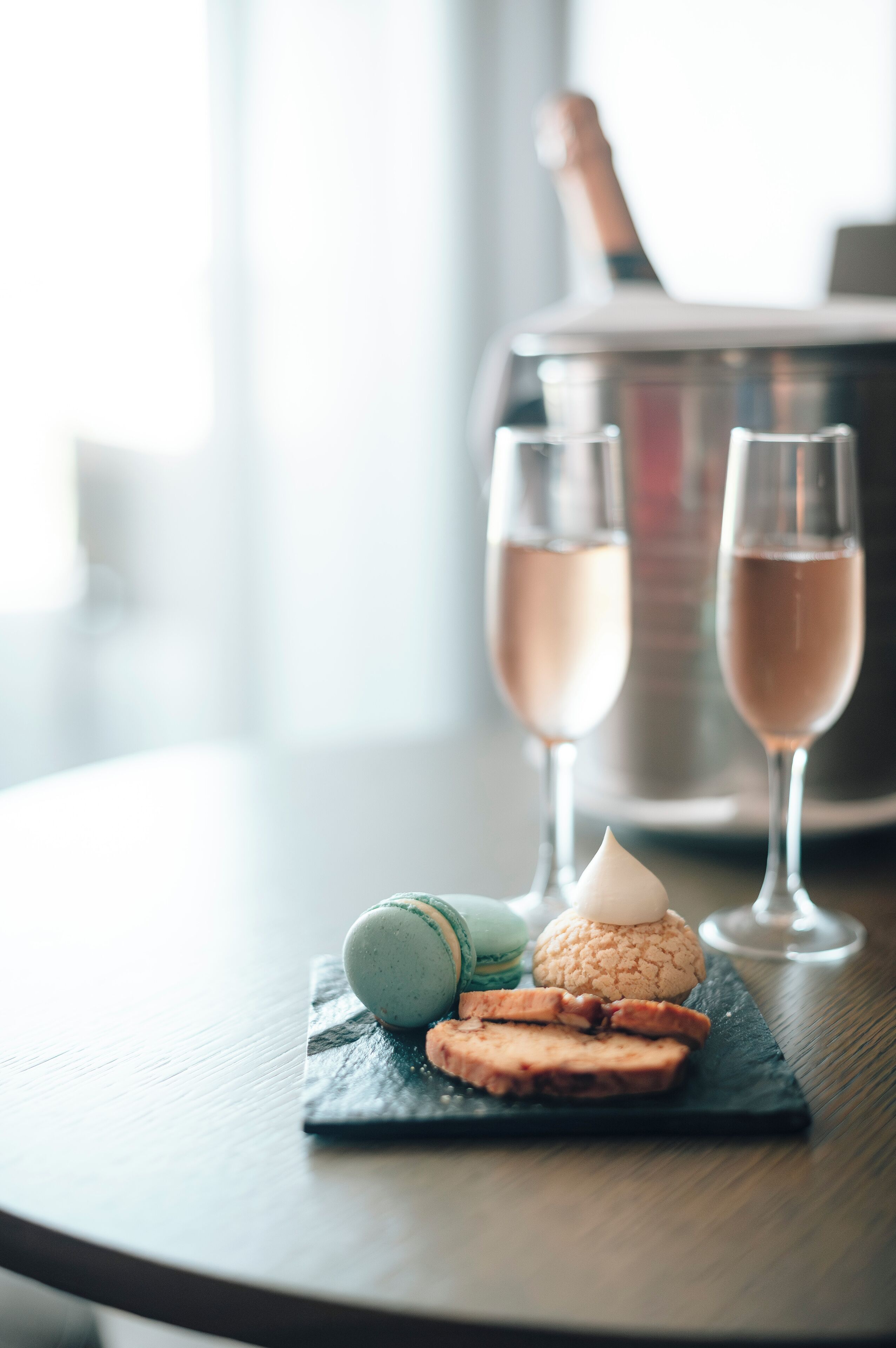 A table with a dessert plate containing macarons and cookies, two glasses of champagne, and a bottle in the background.
